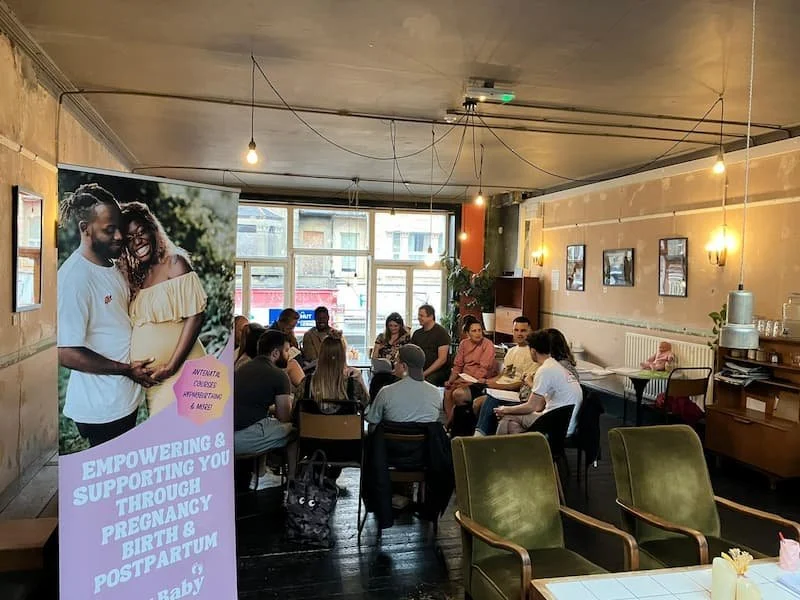 Participants sat in a circle on chairs at an antenatal class with a sign in the foreground that says 'empowering & supporting you through pregnancy birth & postpartum'