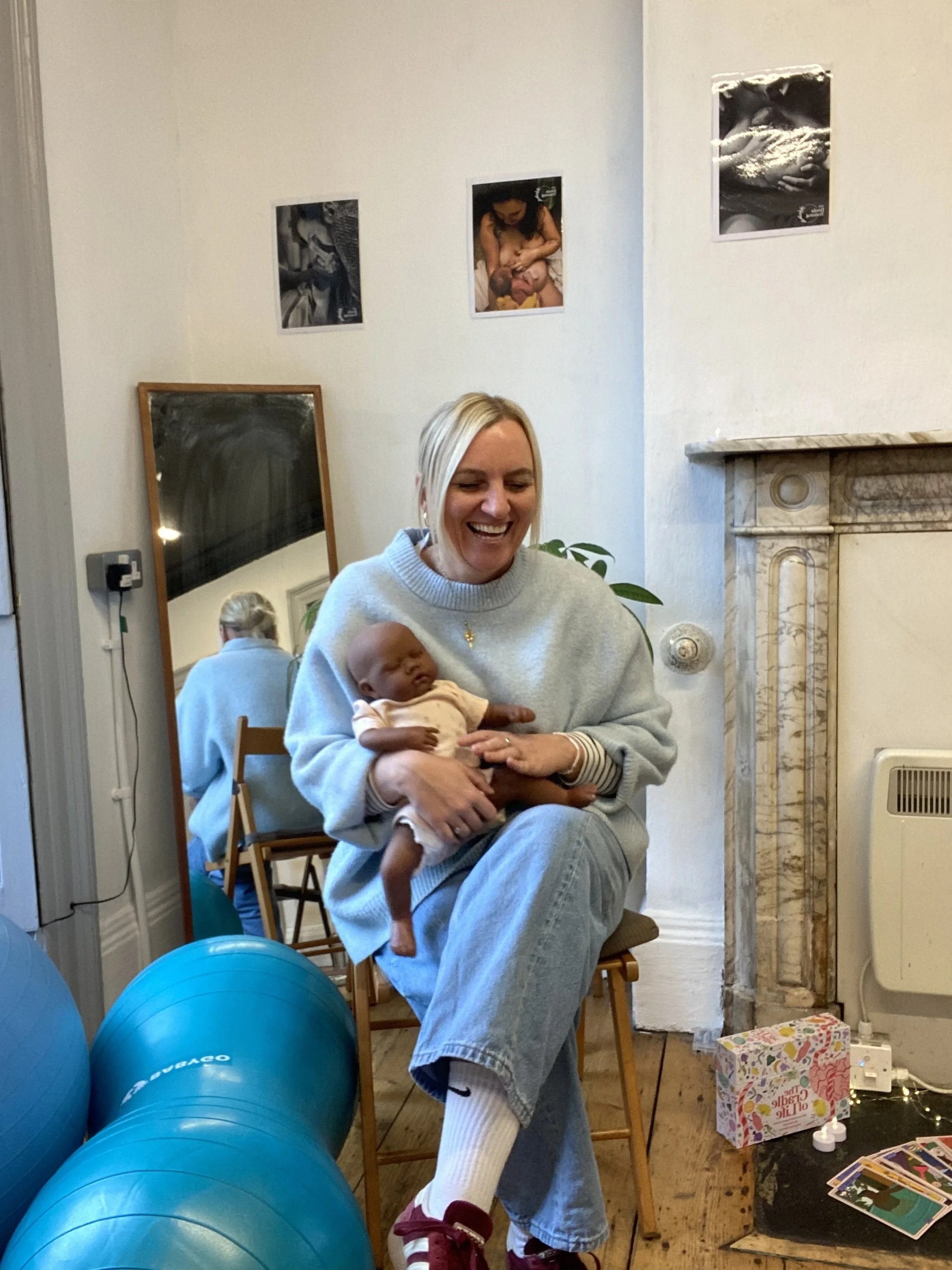 A woman sitting on a wooden chair holding a baby in a cozy room with light-colored walls. There are two blue exercise balls on the floor, a mirror reflecting her back, and framed photos or ultrasound images on the wall. A fireplace mantel and a gift box with colorful wrapping paper are also visible.