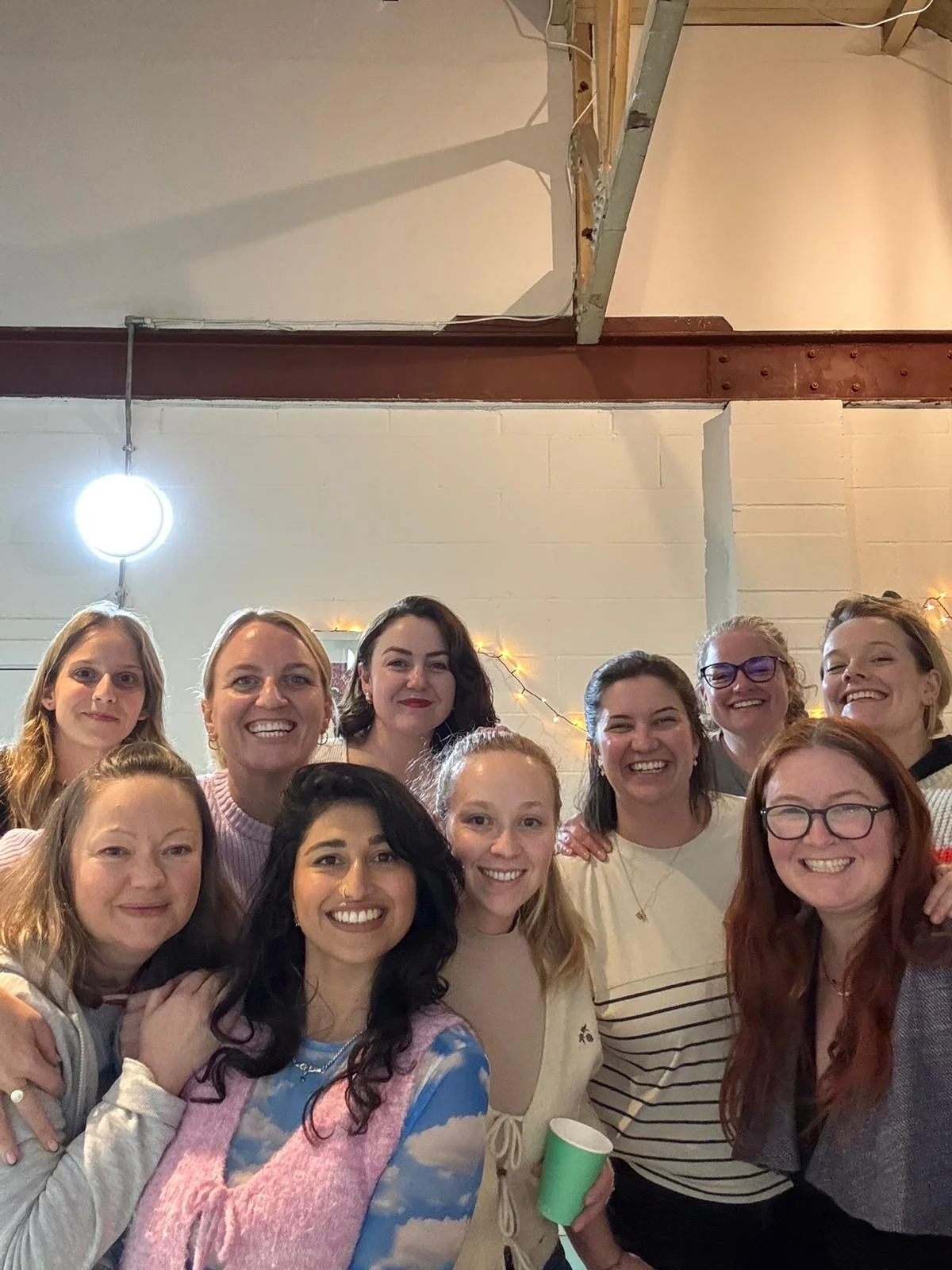 A group of women smiling and posing for a photo at an indoor gathering or party, with string lights and a white wall in the background.