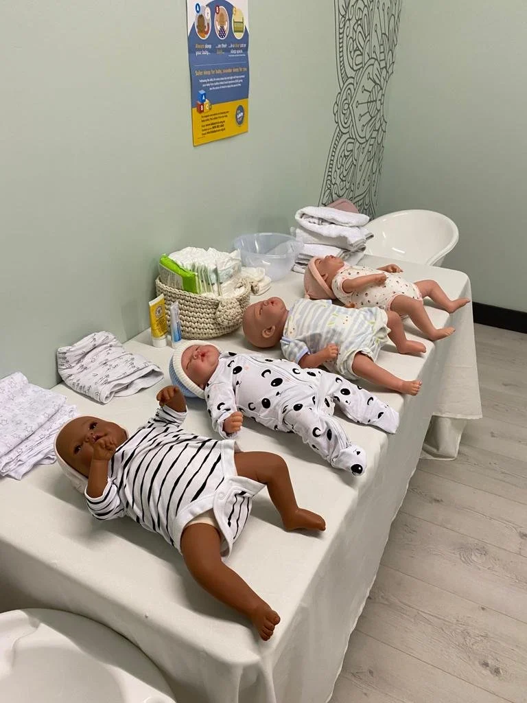 Display of six baby trainng dolls lying on a white changing table under a mint green wall with a colorful poster and a decorative wall pattern.