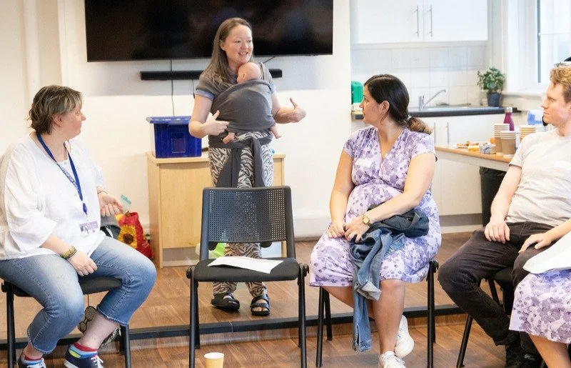 A woman holding a baby, standing and smiling during a group meeting in a room with four seated adults, some wearing hospital scrubs, with a TV mounted on the wall behind.