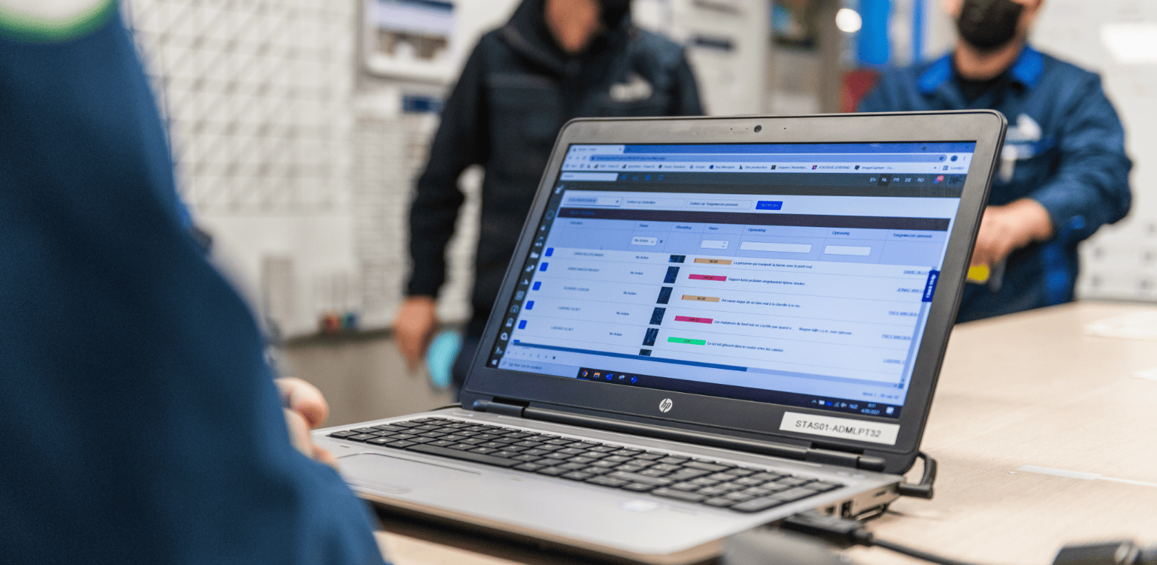 A person using a laptop with a spreadsheet open in a work environment, blurred colleagues in the background.