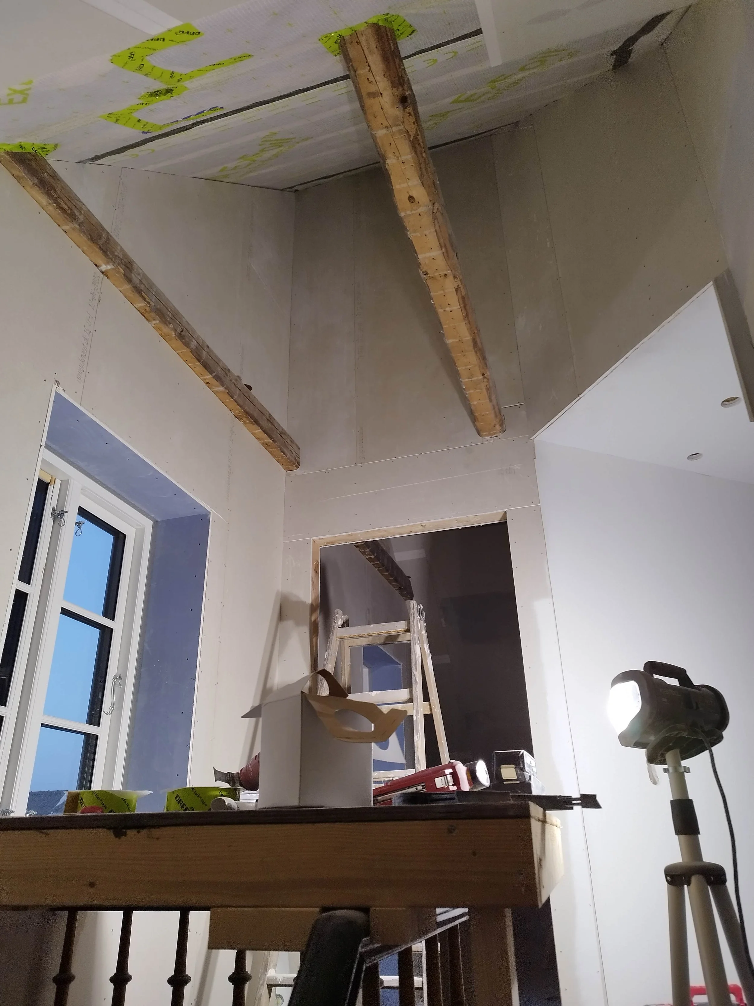 Værelset under renovering med synlige loftsbjælker, arbejdsbord med værktøj, stige i baggrunden og en arbejdslygte på stativ.