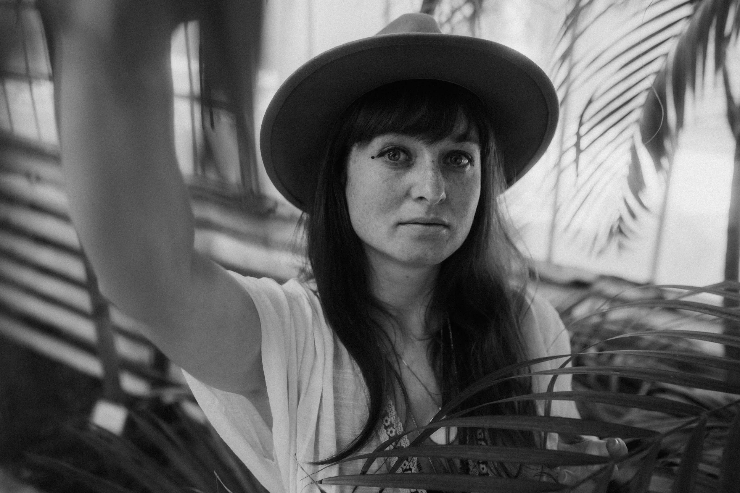 A black and white photograph of a woman with long dark hair and freckles, wearing a wide-brimmed hat and looking directly at the camera, surrounded by palm leaves.