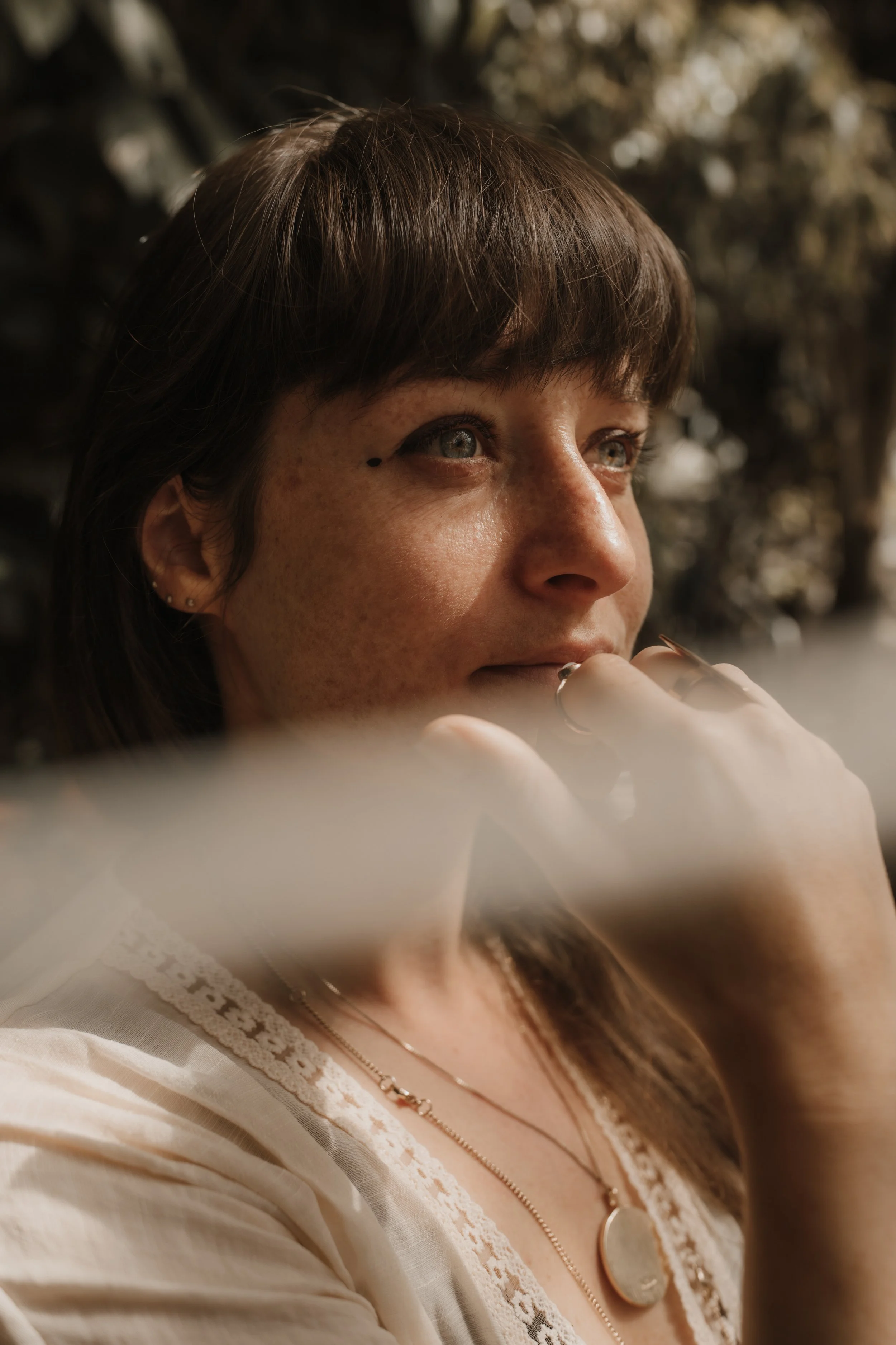 Close-up of a woman with brown hair and blue eyes, wearing a white top with lace detailing and layered necklaces, with a blurred background.