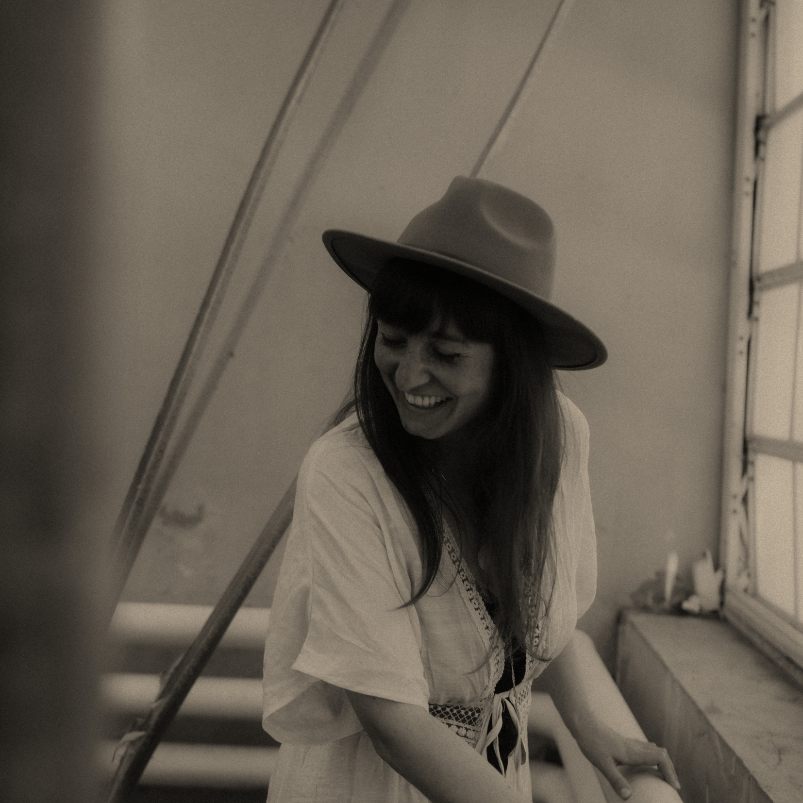 A woman smiling, wearing a wide-brimmed hat and a light-colored blouse, standing beside a diagonal structure in an indoor setting.