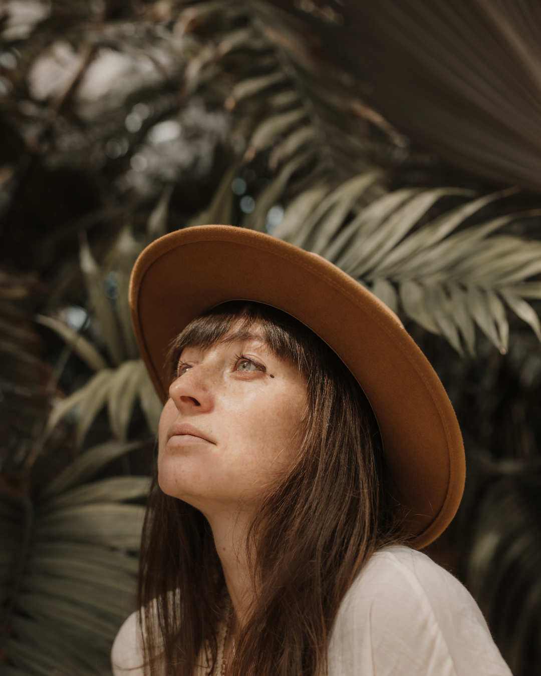 A woman with long brown hair wearing a wide-brimmed brown hat, looking upward, in front of tropical plants.