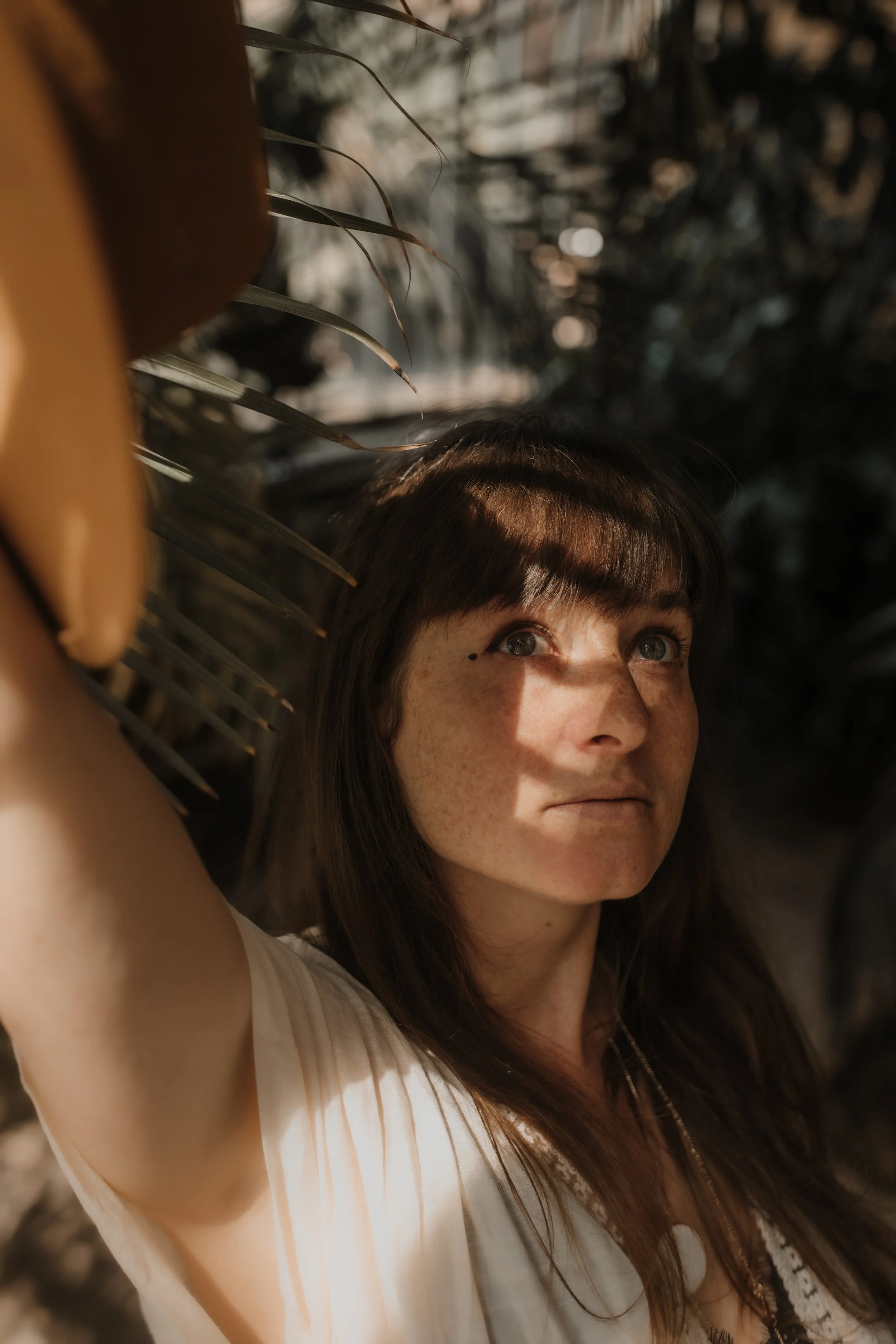 A woman with brown hair and blue eyes taking a selfie outdoors, with a blurred natural background and sunlight on her face.