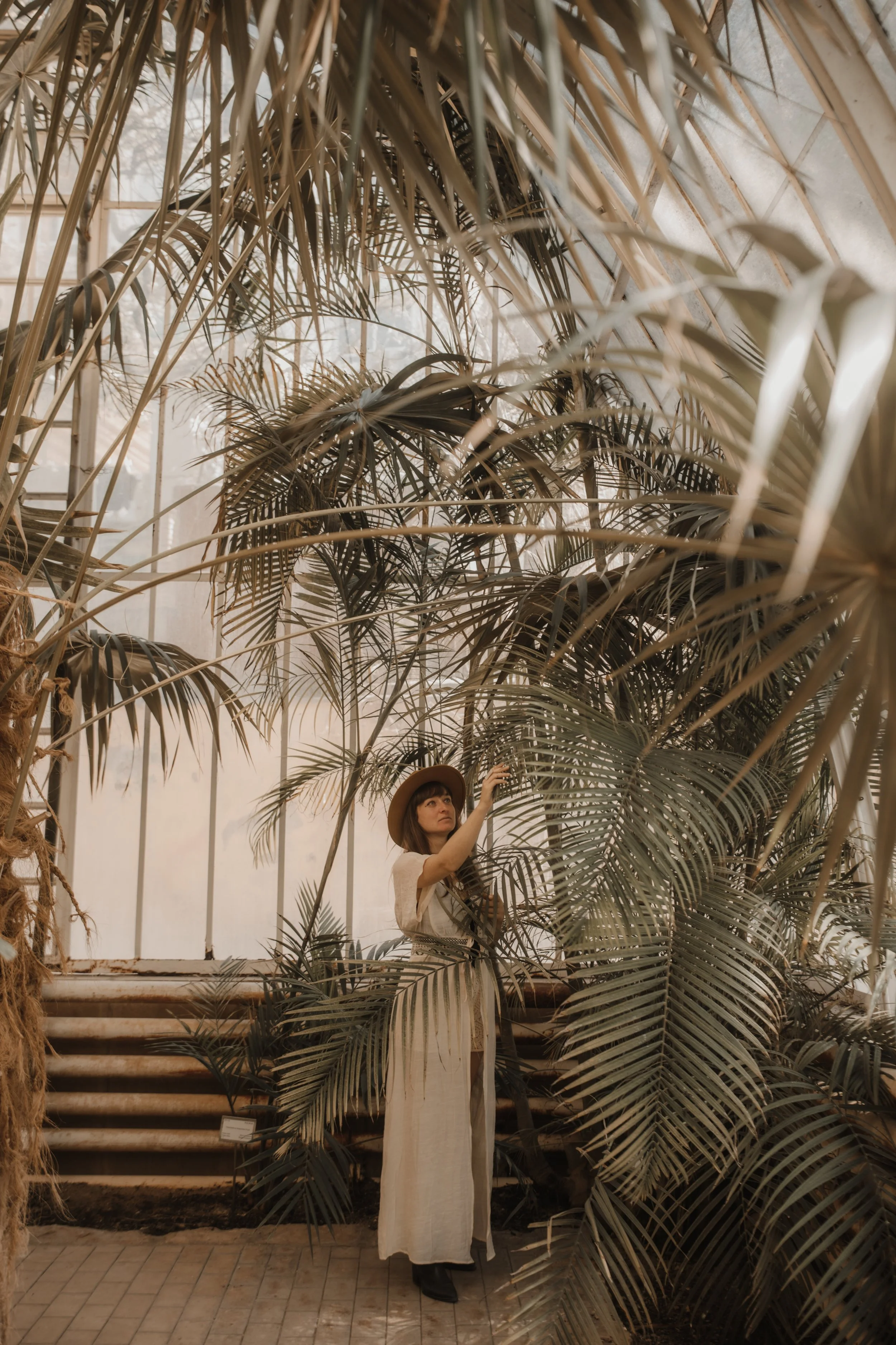 Woman in a long, light-colored dress and wide-brimmed hat standing amidst lush, large leafy tropical plants inside a glass greenhouse.