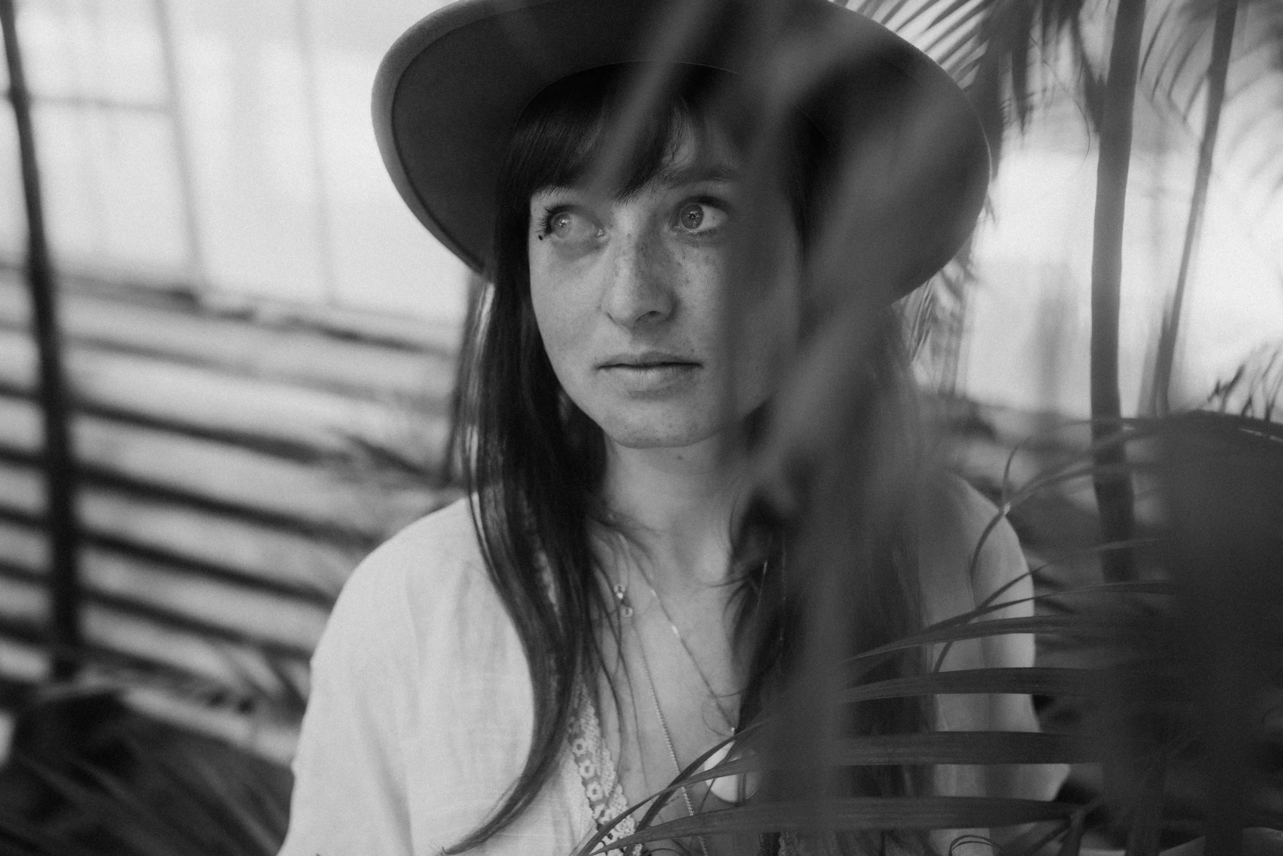 Schwarz-weiß-Foto eines mädchen mit langen dunklen haaren, getragen einem Hut und einer Bluse, umgeben von Pflanzen.