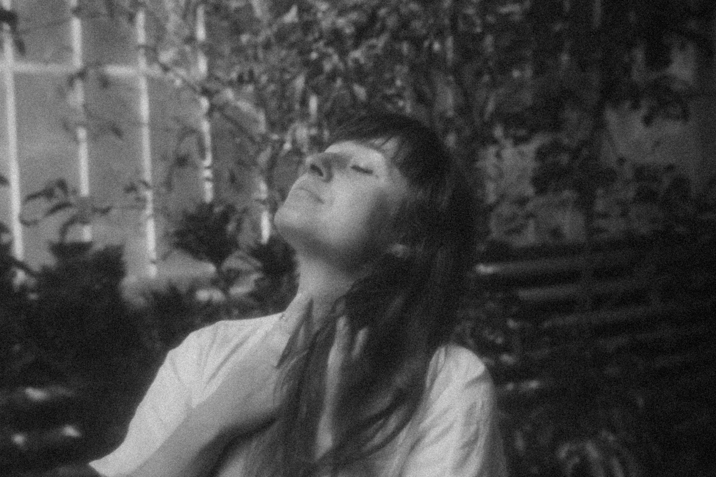 A woman with long dark hair and closed eyes standing outdoors in front of a fence and leafy background.
