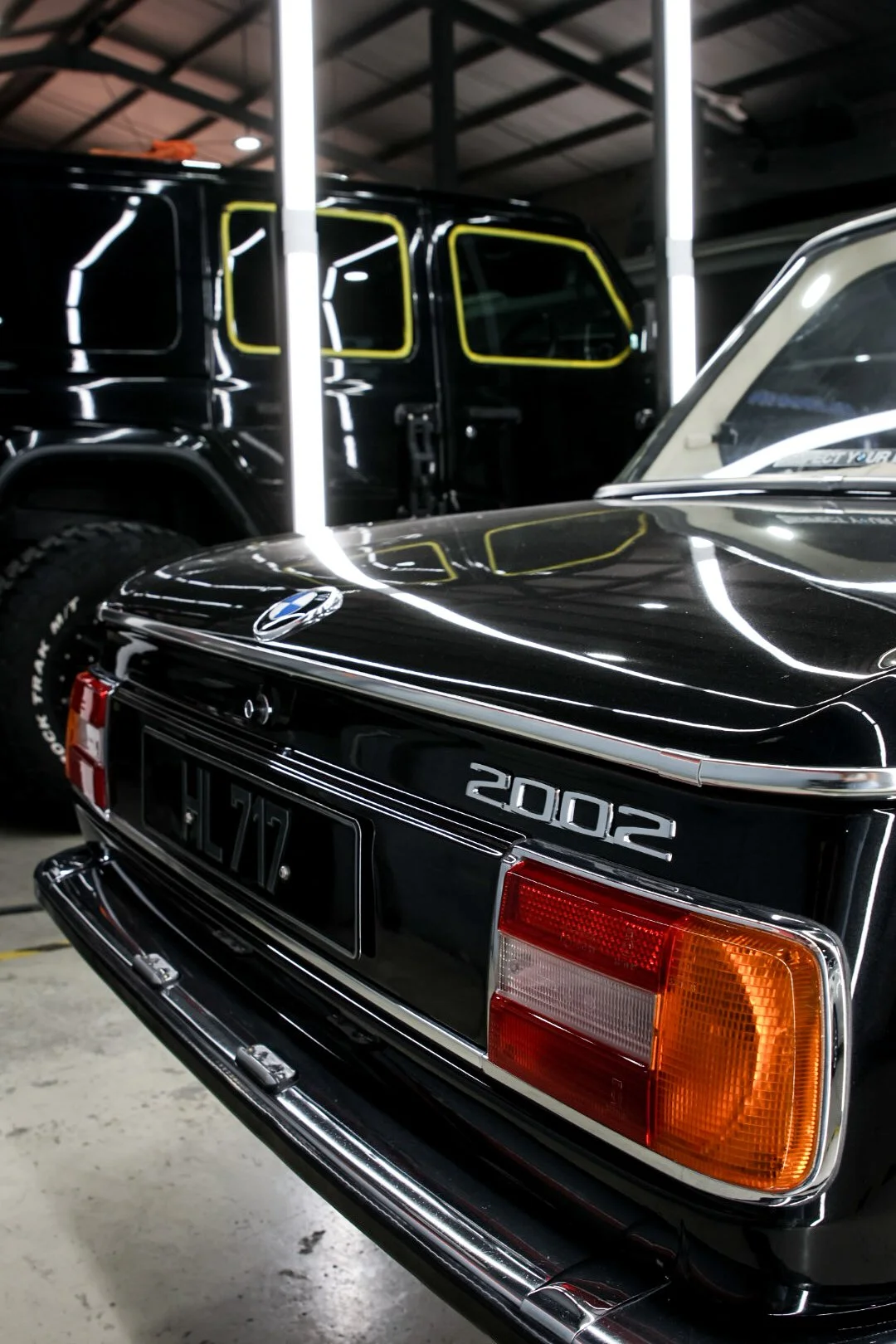 The rear of a vintage black BMW 2002 car, showing the model badge and a distinctive tail light, parked inside a garage or showroom with fluorescent lighting, reflection of the lights on the car's surface, and a black vehicle with yellow outlines of windows in the background.