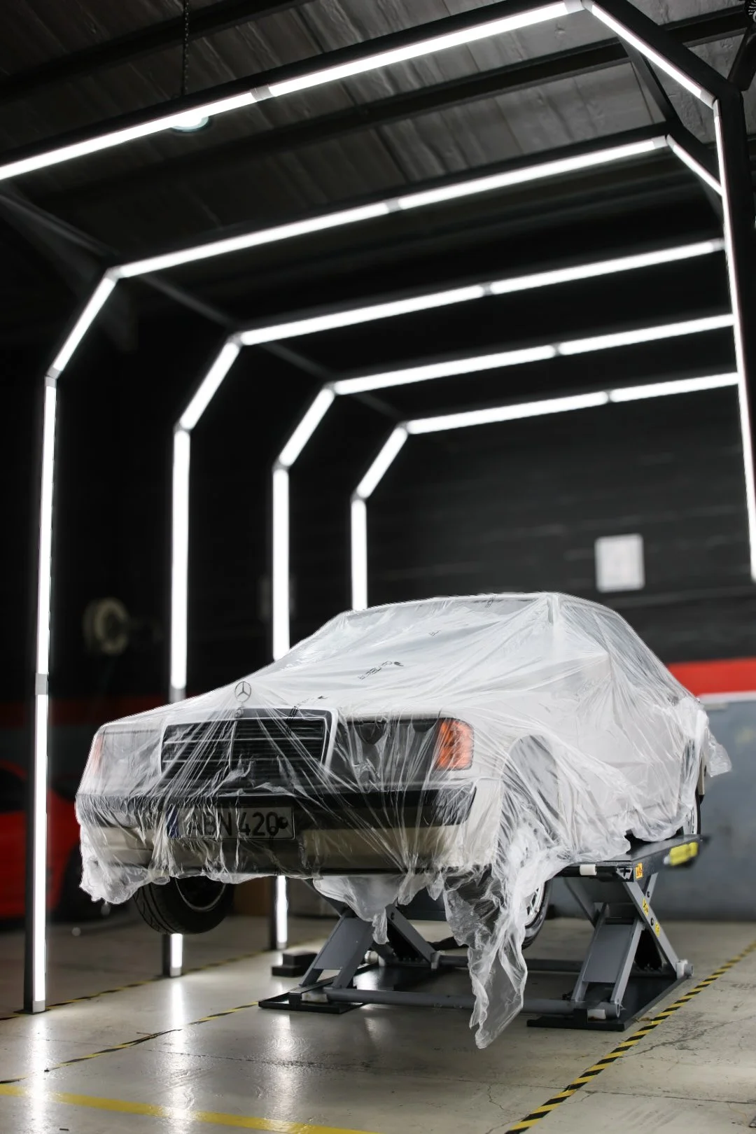 A classic white Mercedes-Benz car partially wrapped in protective plastic, elevated on a scissor lift in a garage or workshop with black walls and white LED strip lighting.