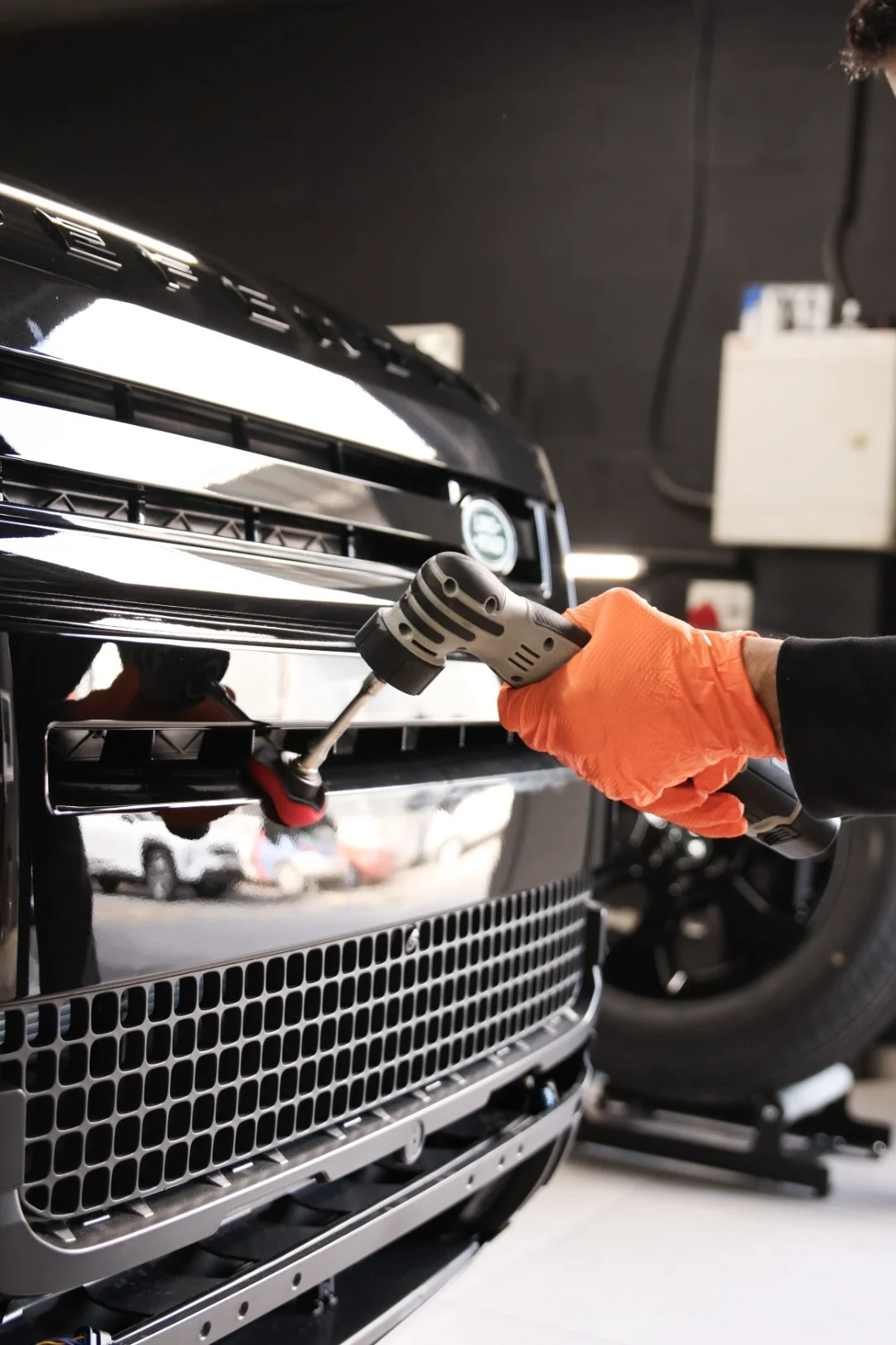 A person wearing orange gloves using a drill to work on the front grille of a black luxury car, with other cars and tools visible in the background.