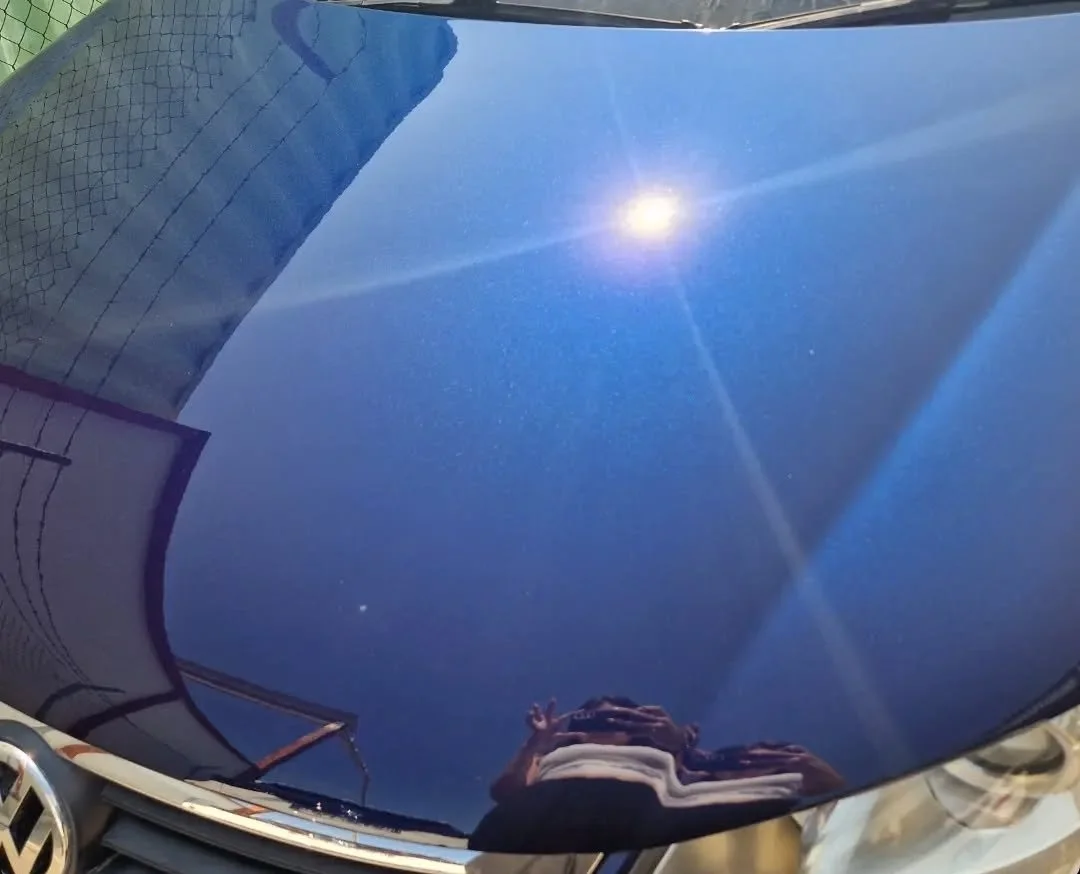 Reflection of the blue sky with the bright sun and some buildings on the shiny black surface of a car hood.