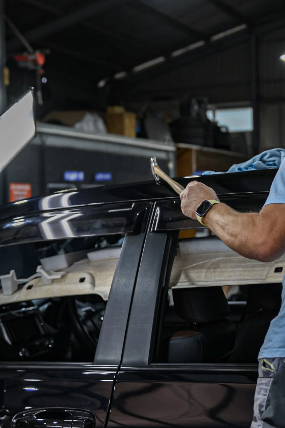 Person working on a black car in a garage, using a tool to adjust the car's roof or trim, with a smartwatch on their left wrist.