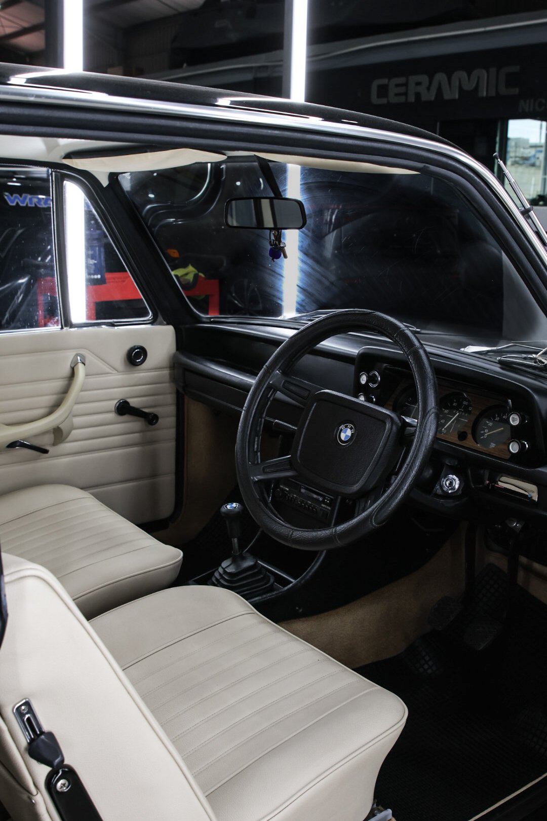 Interior of a vintage BMW car, showing the steering wheel, black dashboard, beige seats and door panel, gear shift, and rearview mirror.