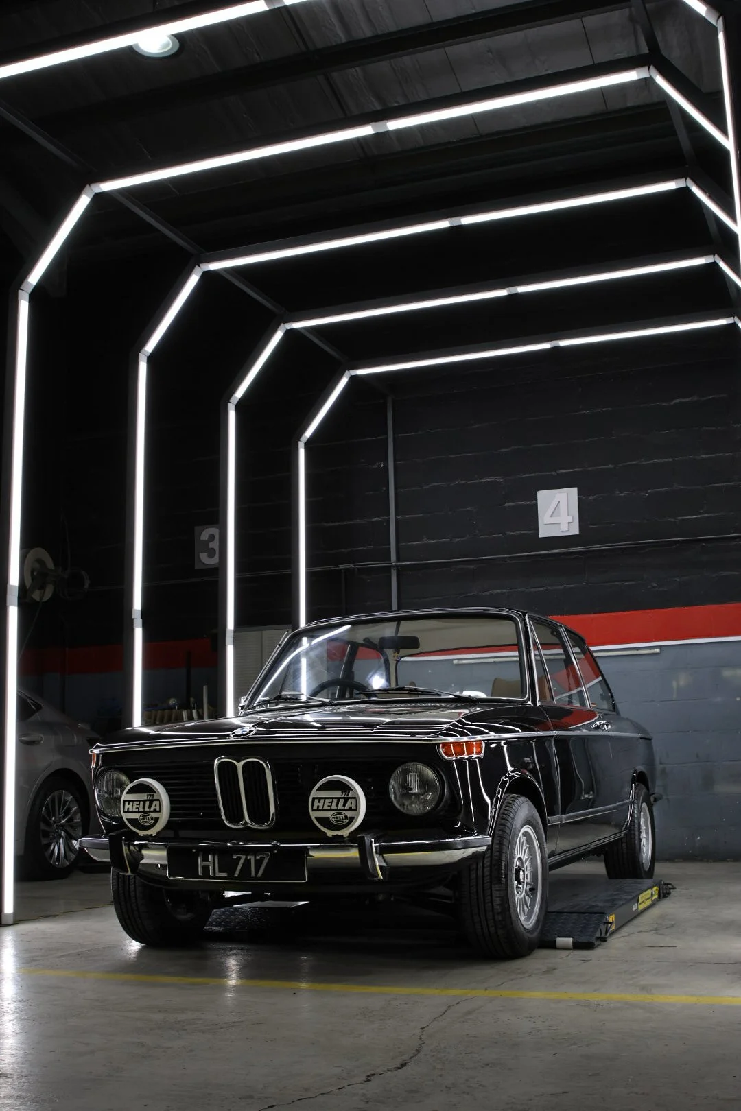 A vintage black car with rally lights in an indoor garage with modern LED lighting and numbered sections.