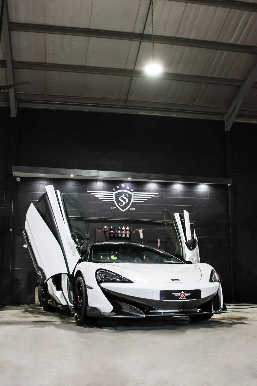 White sports car with gull-wing doors open inside a garage with black walls and a logo on the wall that reads 'EST 1998' and features a shield, wings, and stars.