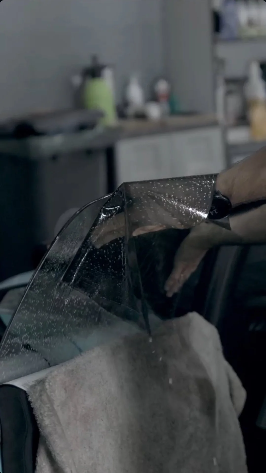 A person is installing a clear protective shield on a vehicle, with a kitchen or workshop in the background.