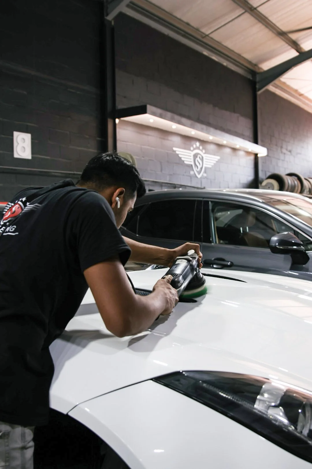 A man polishing the hood of a white sports car with an electric orbital buffer in an indoor garage, with a black car parked behind him and a logo on the wall.