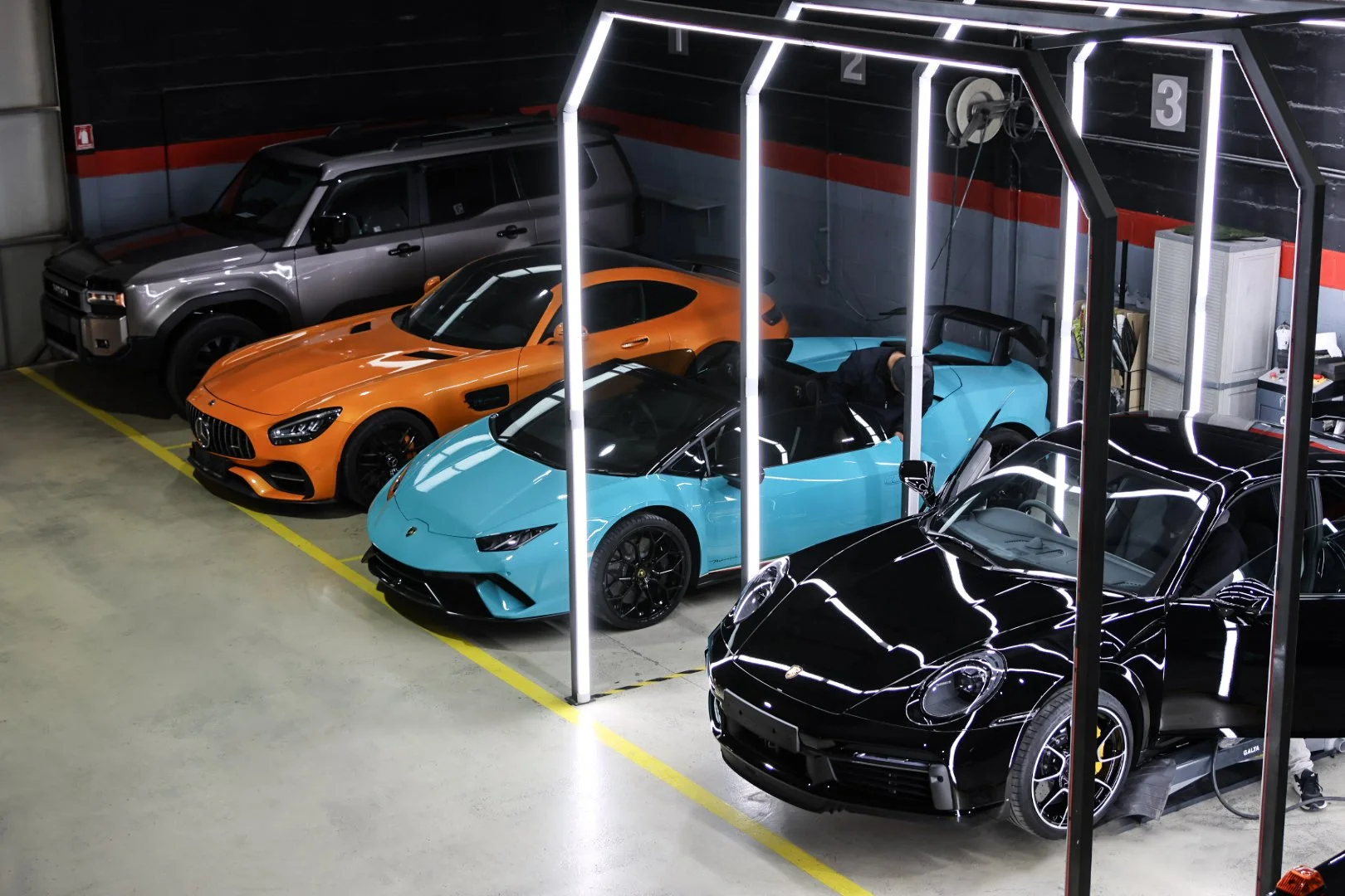 A row of luxury cars parked in a garage, including a black Porsche, a blue Lamborghini, an orange Mercedes-Benz AMG, and a gray Land Rover, with bright vertical LED lights.