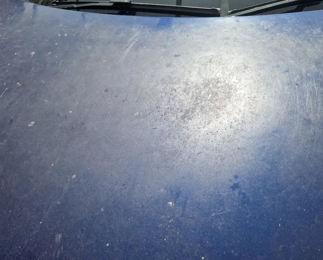 Close-up of a dark blue car hood with visible scratches, dirt, and a reflection of sunlight.