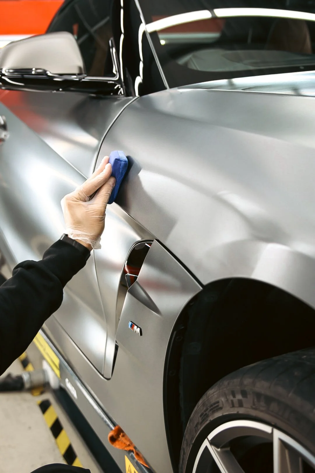 Person wearing black gloves polishing the silver body of a sports car with a blue cloth in an indoor garage.