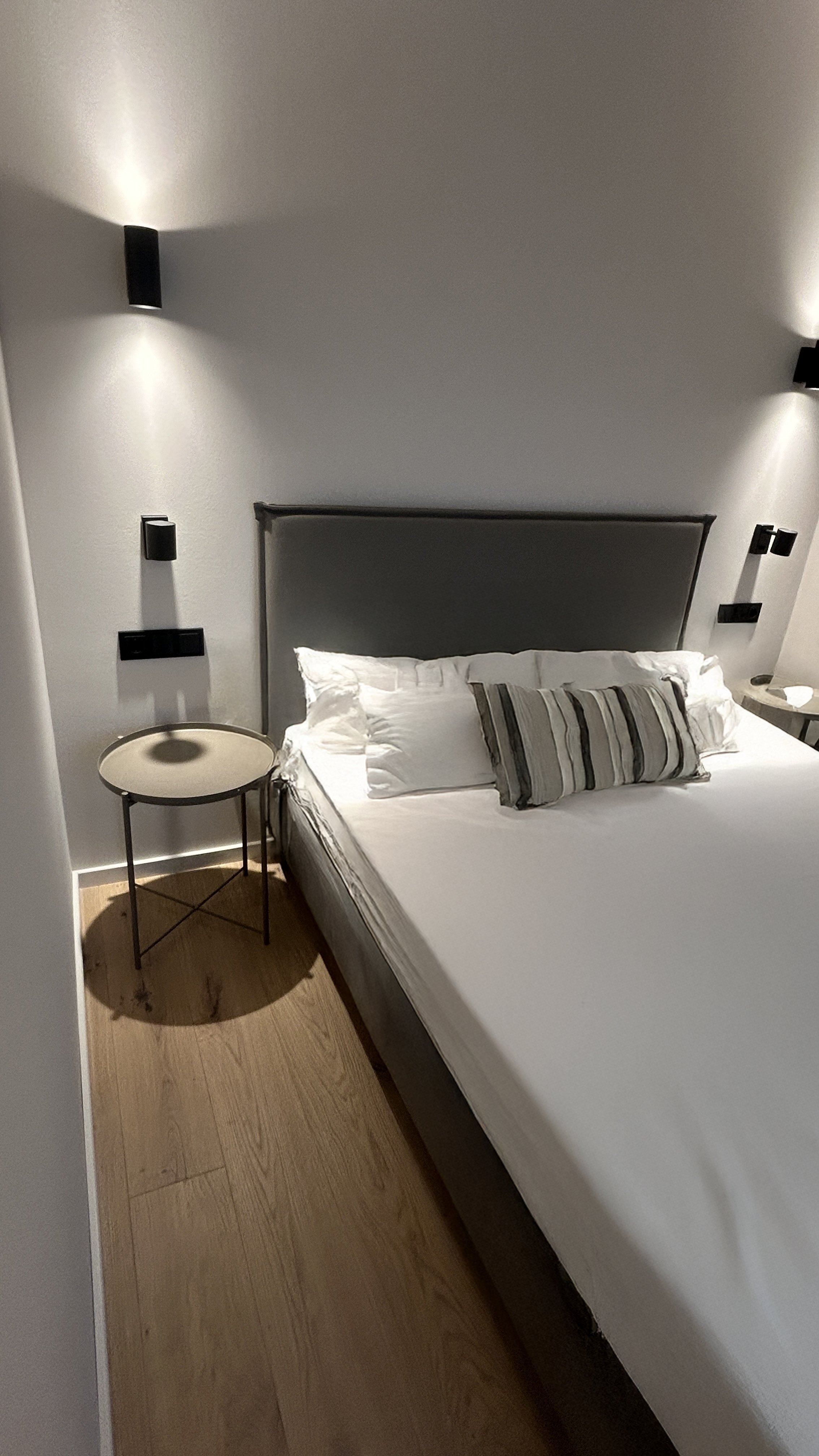 Modernes Schlafzimmer mit großem Bett, weißen Kissen und gestreiftem Zierkissen, Holzfußboden, runder Nachttisch, schlichte Wandleuchten an weißer Wand.