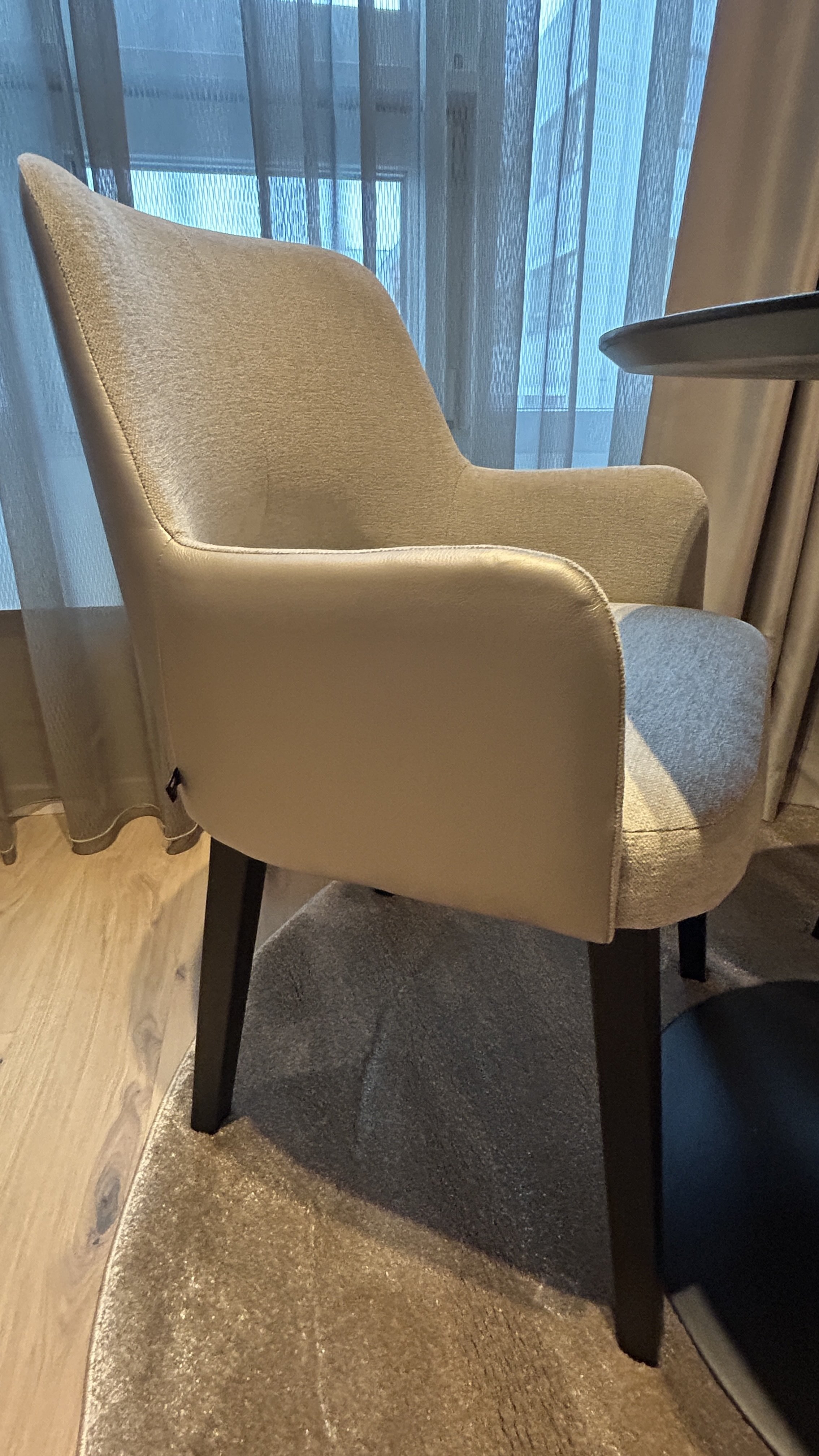 Grau gepolsterter Stuhl mit Armlehnen, stehend auf einem Teppich, vor einem Fenster mit durchsichtigen Vorhängen.