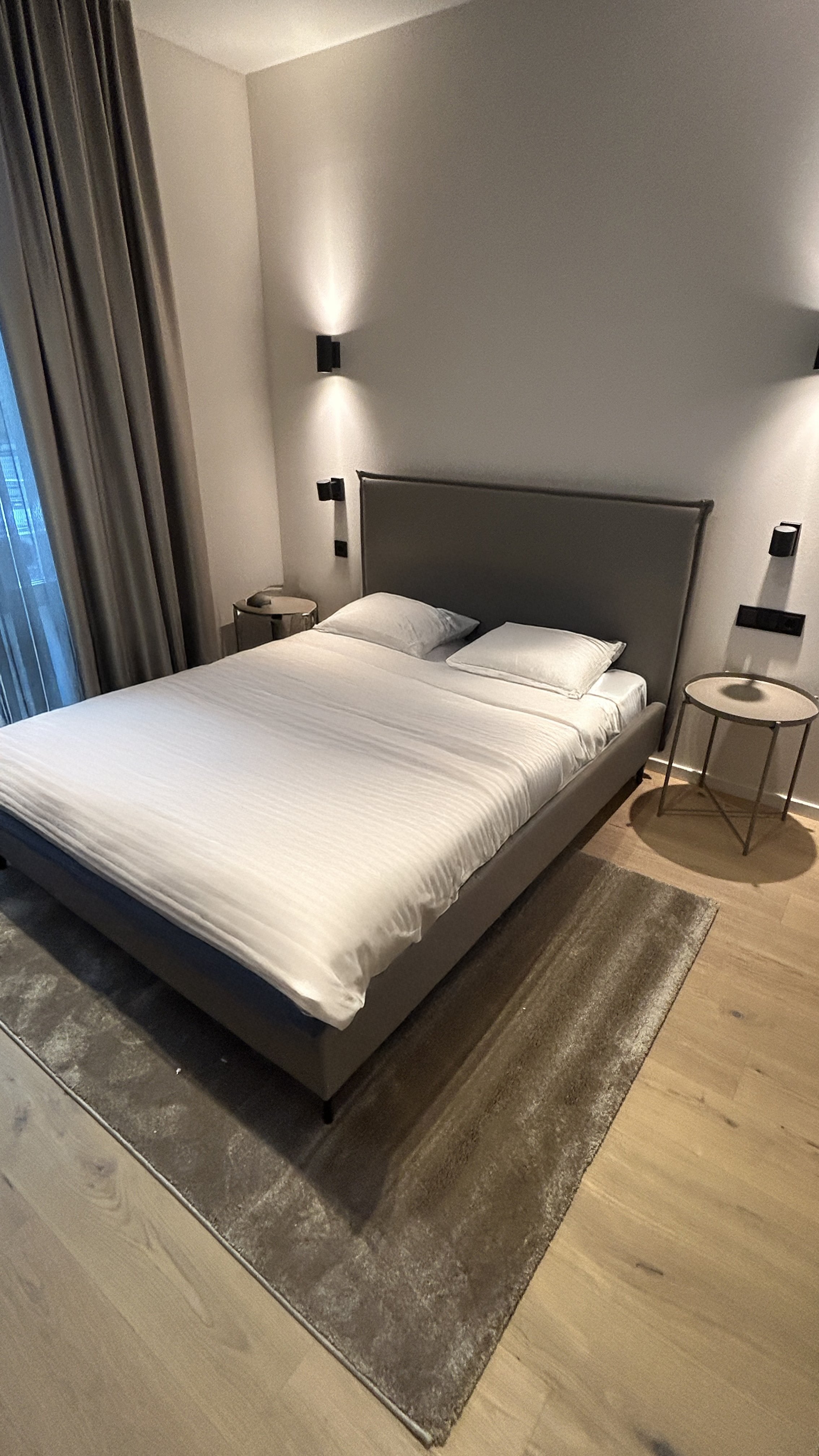 Minimalistisch eingerichtetes Schlafzimmer mit einem Doppelbett, weißen Bettbezügen und zwei Nachttischen, auf einem Teppichboden, beleuchtet von Wandlampen.
