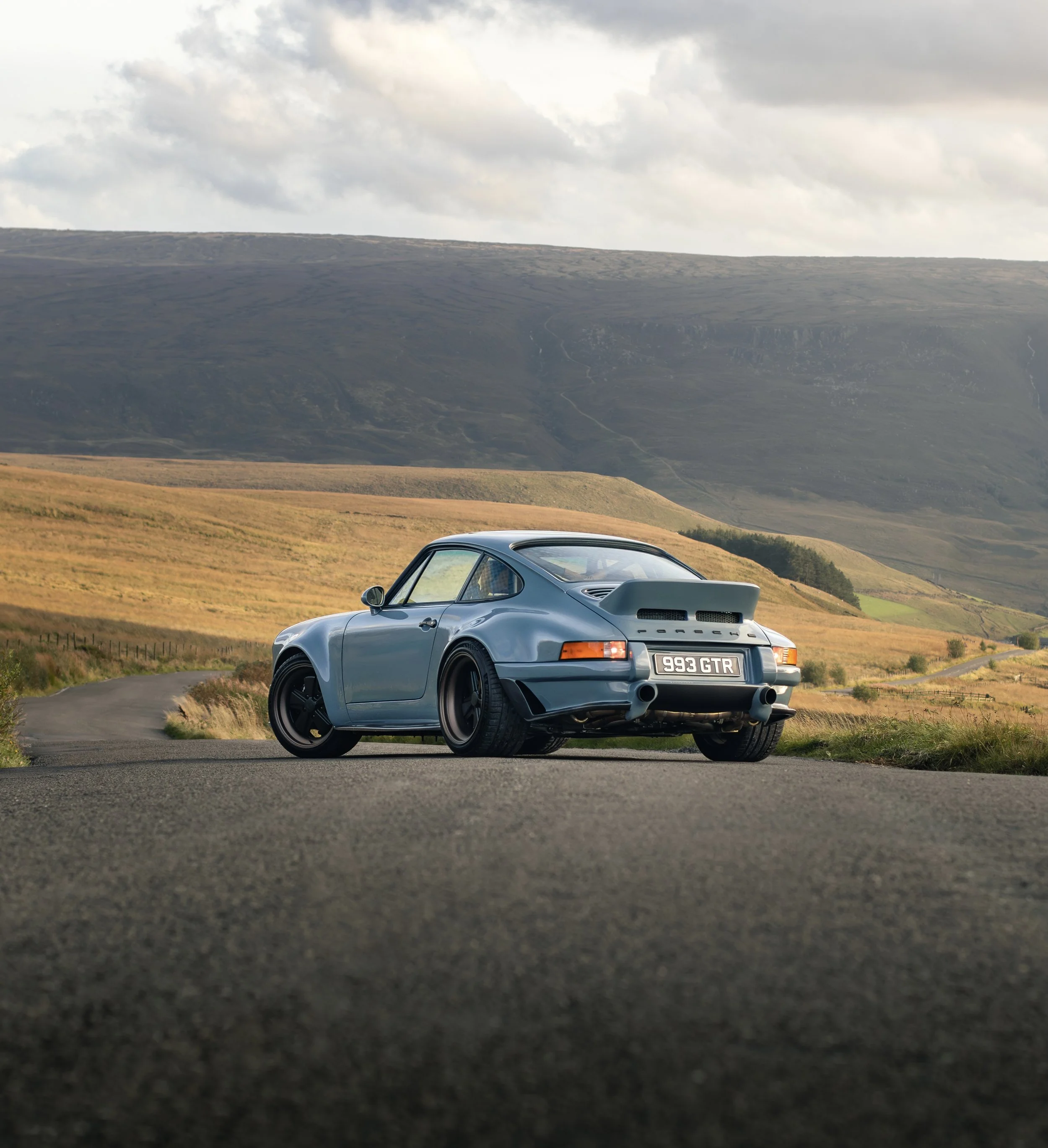 A classic blue Porsche 911 parked on a winding road in a rural landscape with rolling hills and a cloudy sky in the background.