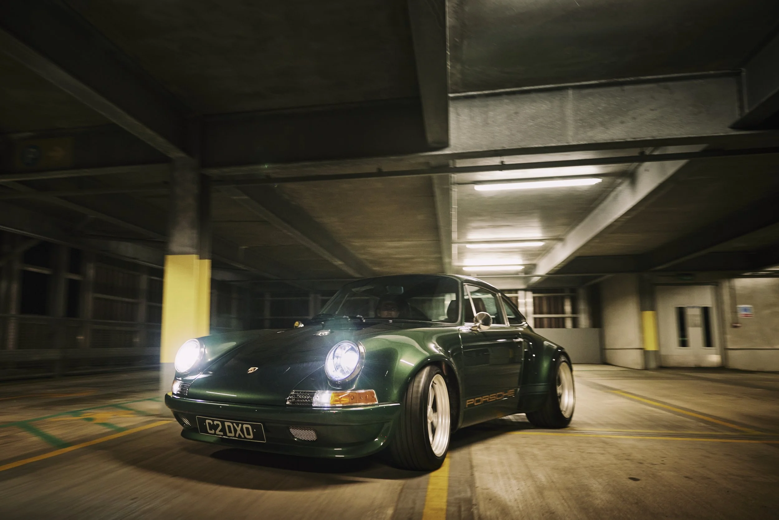 A dark green vintage Porsche 911 Carrera in an empty parking garage at night, with headlights on. The car has a black license plate reading 'C2 DXO' and features a gold 'Porsche' logo on the side.