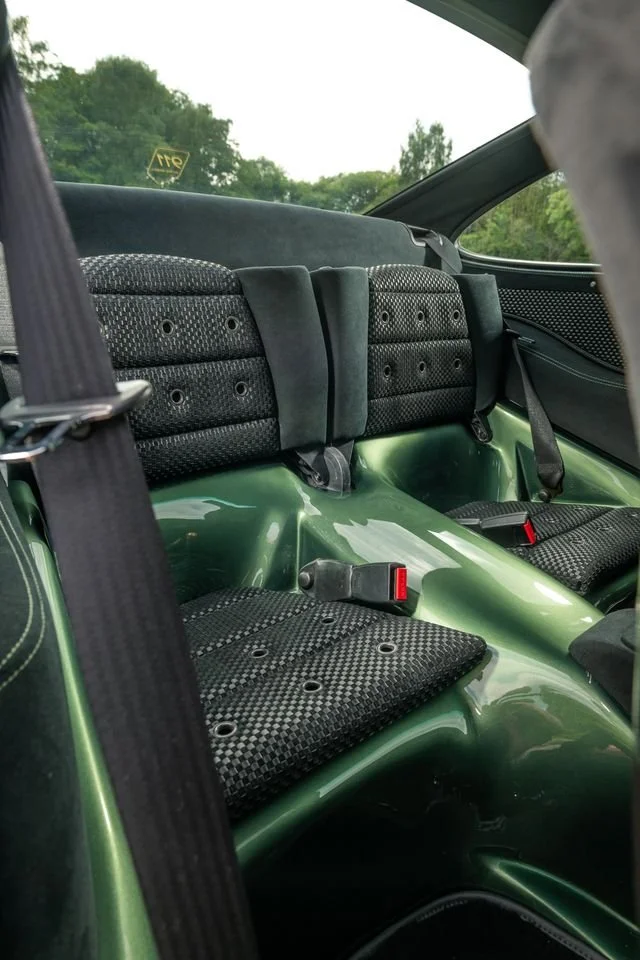 The interior of a vintage green race car, showing two black fabric seats with visible mounting holes and a seatbelt, and a rear window with trees outside.