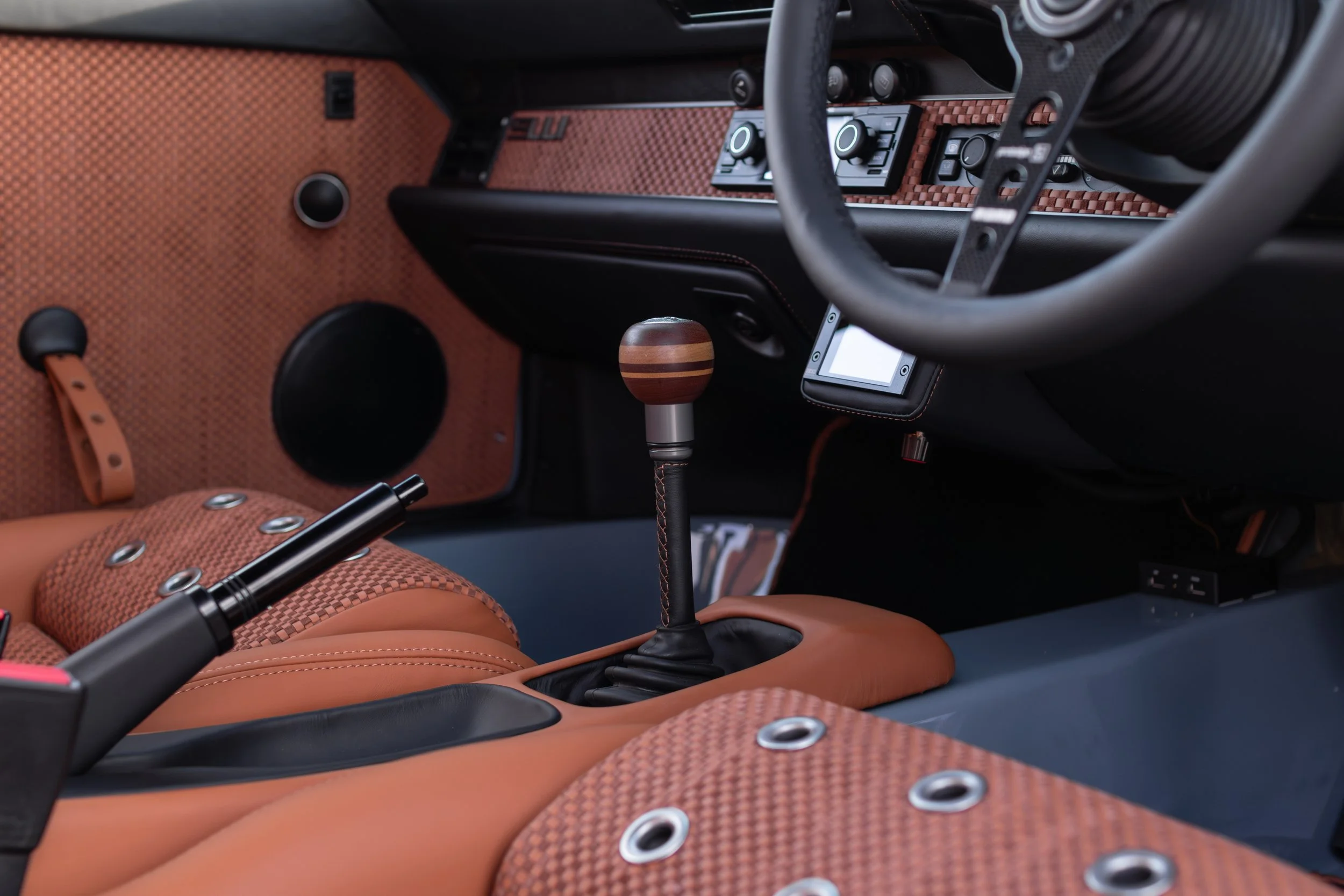 Close-up of a car interior showing a gear shift with a wooden knob, brown and black upholstery with metal grommets, a black steering wheel, and various control buttons on the dashboard.