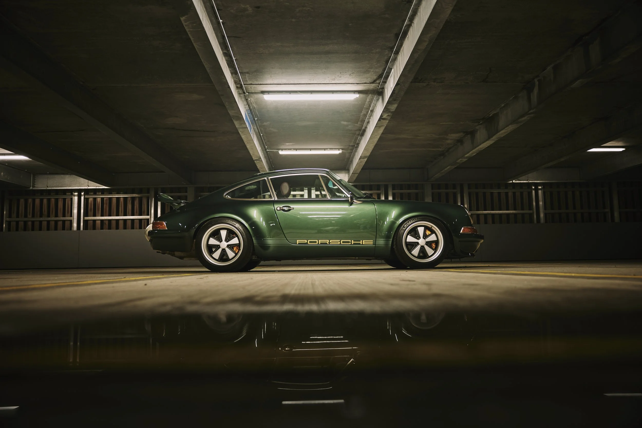 A vintage green Porsche sports car parked in an indoor parking garage with concrete ceiling and yellow parking lot lines.