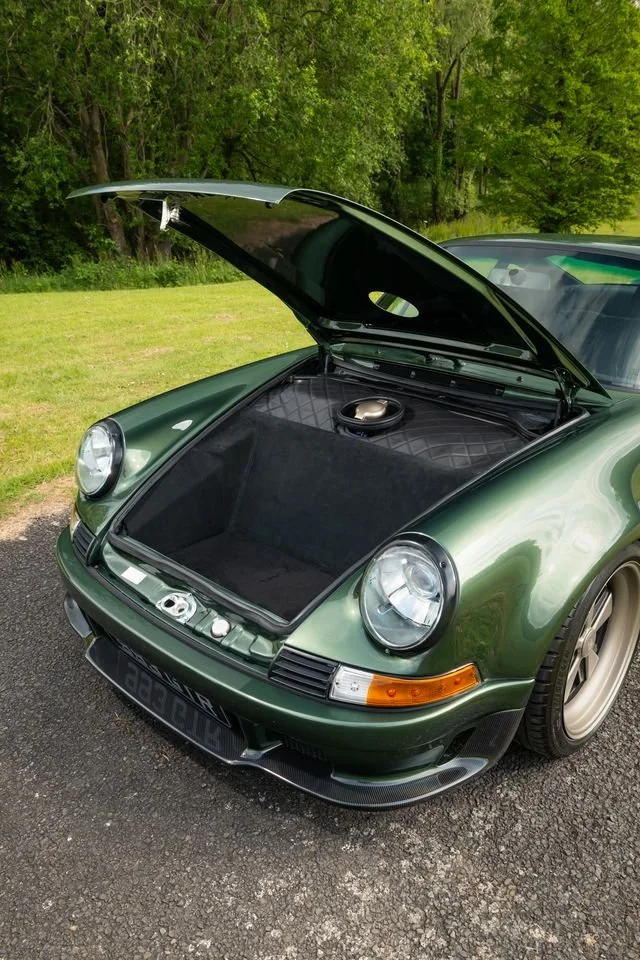 A green vintage Porsche 911 parked on a paved surface with its front trunk open. The trunk is empty except for a speaker and a sound-absorbing mat. The car is surrounded by green trees and grass.