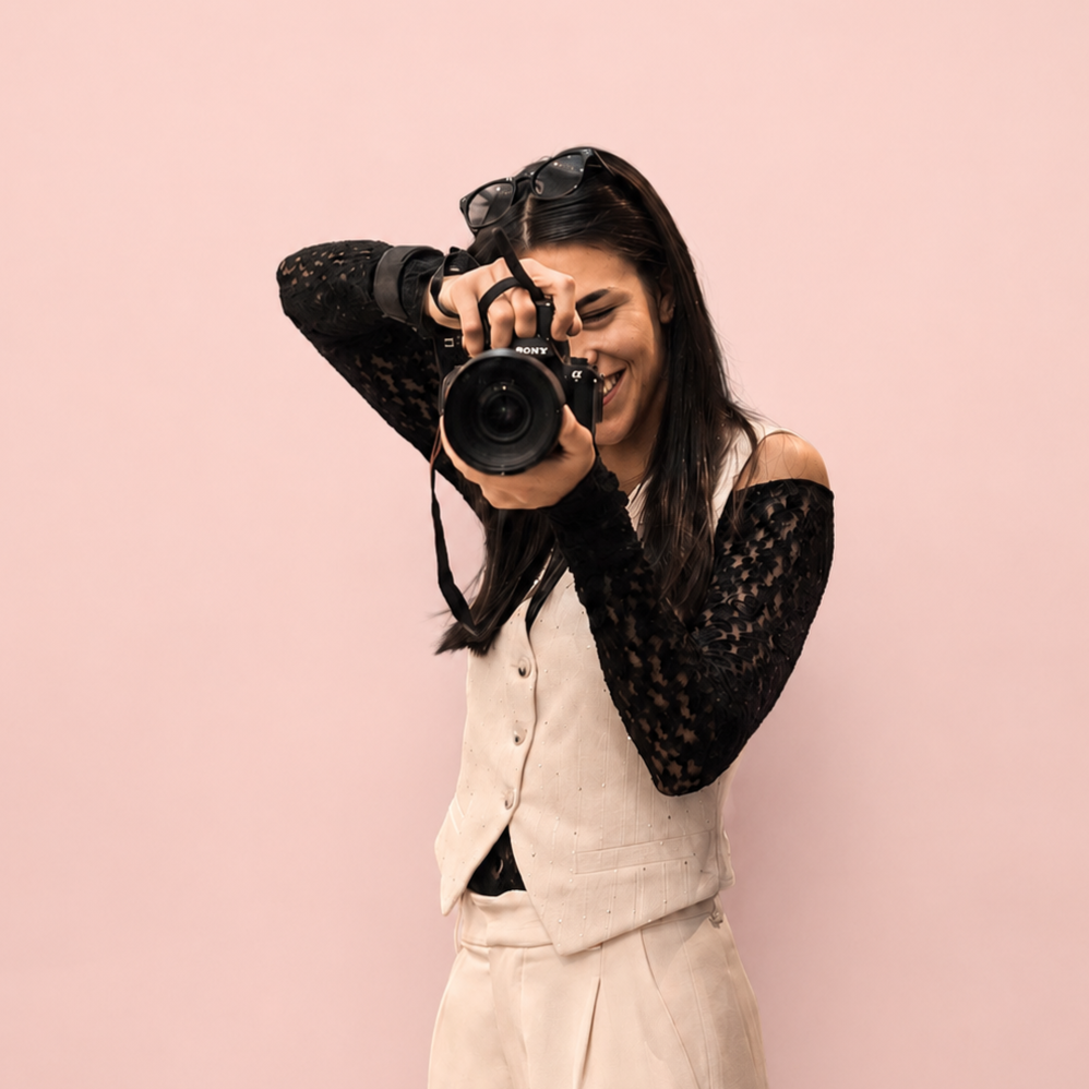 Una donna con camicia beige e giacca nera con dettagli di pizzo, che scatta una foto con una macchina fotografica digitale, sorridendo, con uno sfondo rosa chiaro.