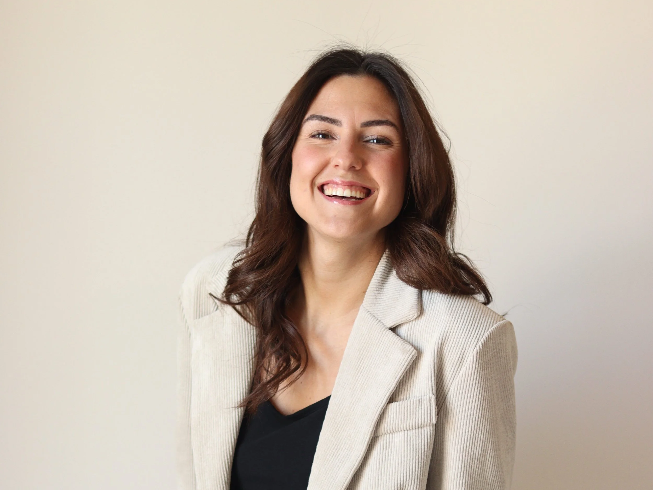 Una donna con capelli castani e riccioli, sorriso amichevole, indossa un cappotto beige e una maglietta nera, sfondo neutro.