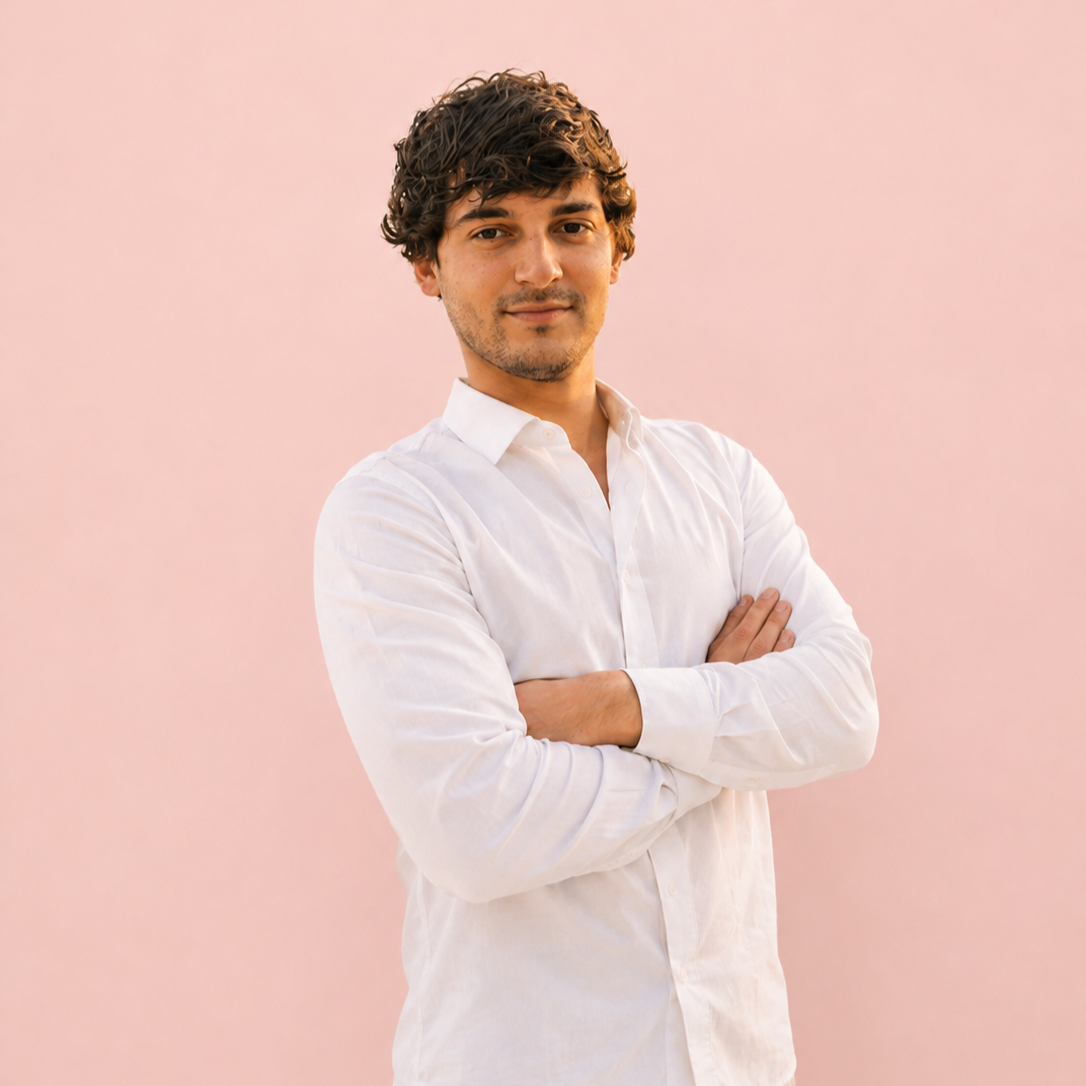 Ragazzo con capelli riccioli scuri indossa camicia bianca, sfondo rosa chiaro.