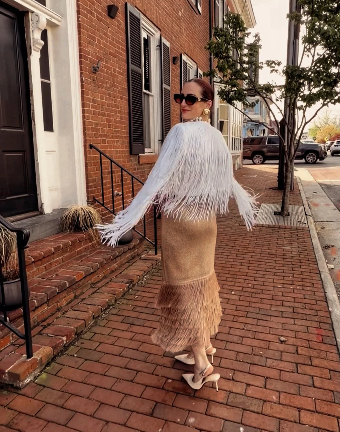 Woman wearing a white feathered jacket, a beige skirt, and high heels, standing on a brick sidewalk in front of a brick building with black shutters, wearing sunglasses, earrings, and a pearl necklace, during daytime.