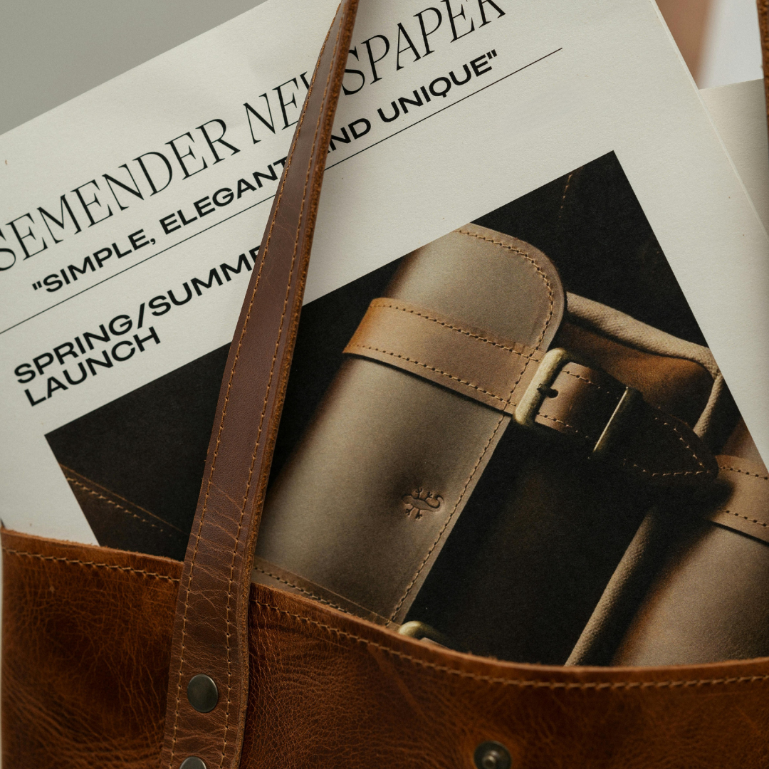A close-up of a magazine inside a brown leather bag, featuring an article about new paper with a photo of a beige strap watch.