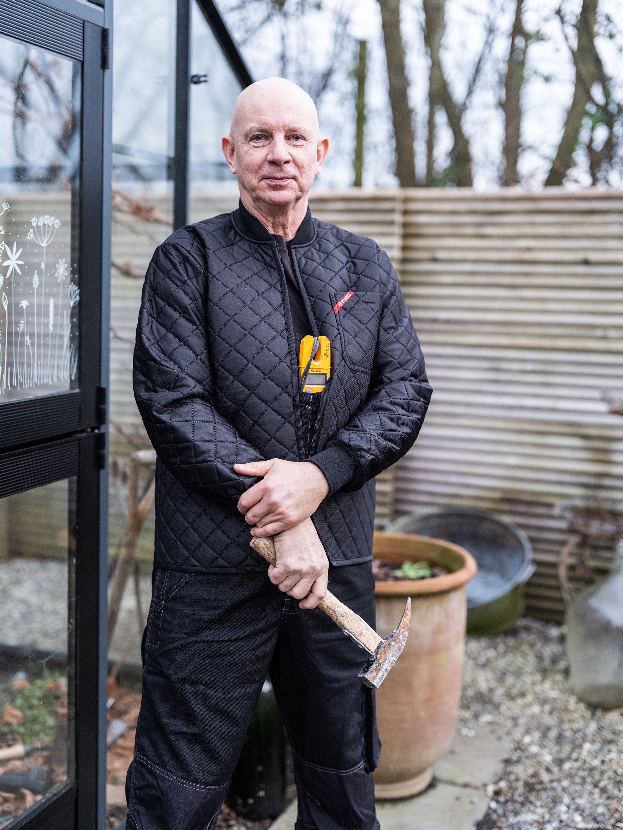 Mand i sort quiltet jakke holder en hammer i en have med blomsterkrukker og glasdør.