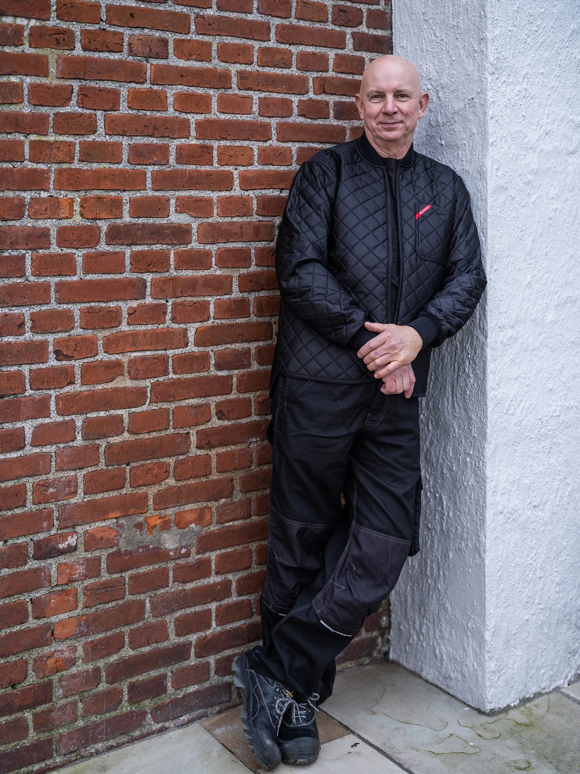 Mand i sort overtøj står ved en murstensvæg og en pudset væg. Han smiler og har hænderne foldet.