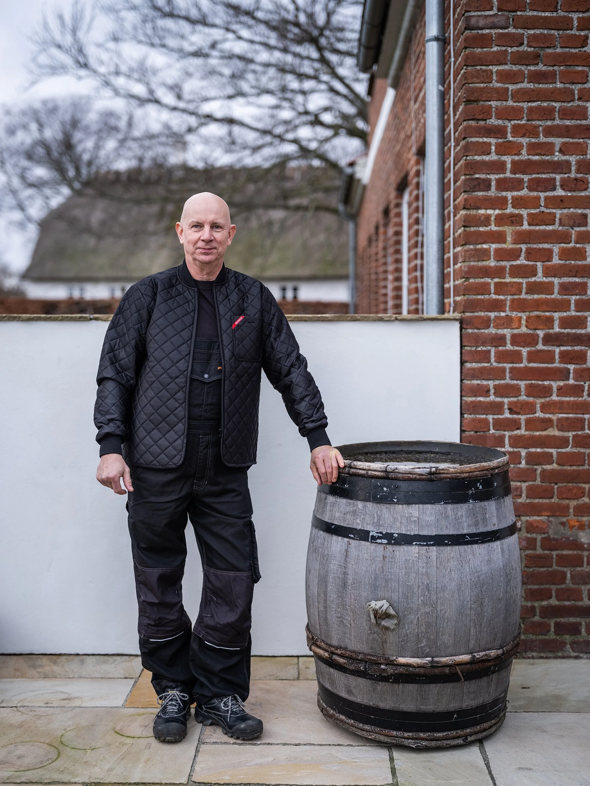 En mand i sort tøj står ved siden af en træfad ude foran en murstensbygning.