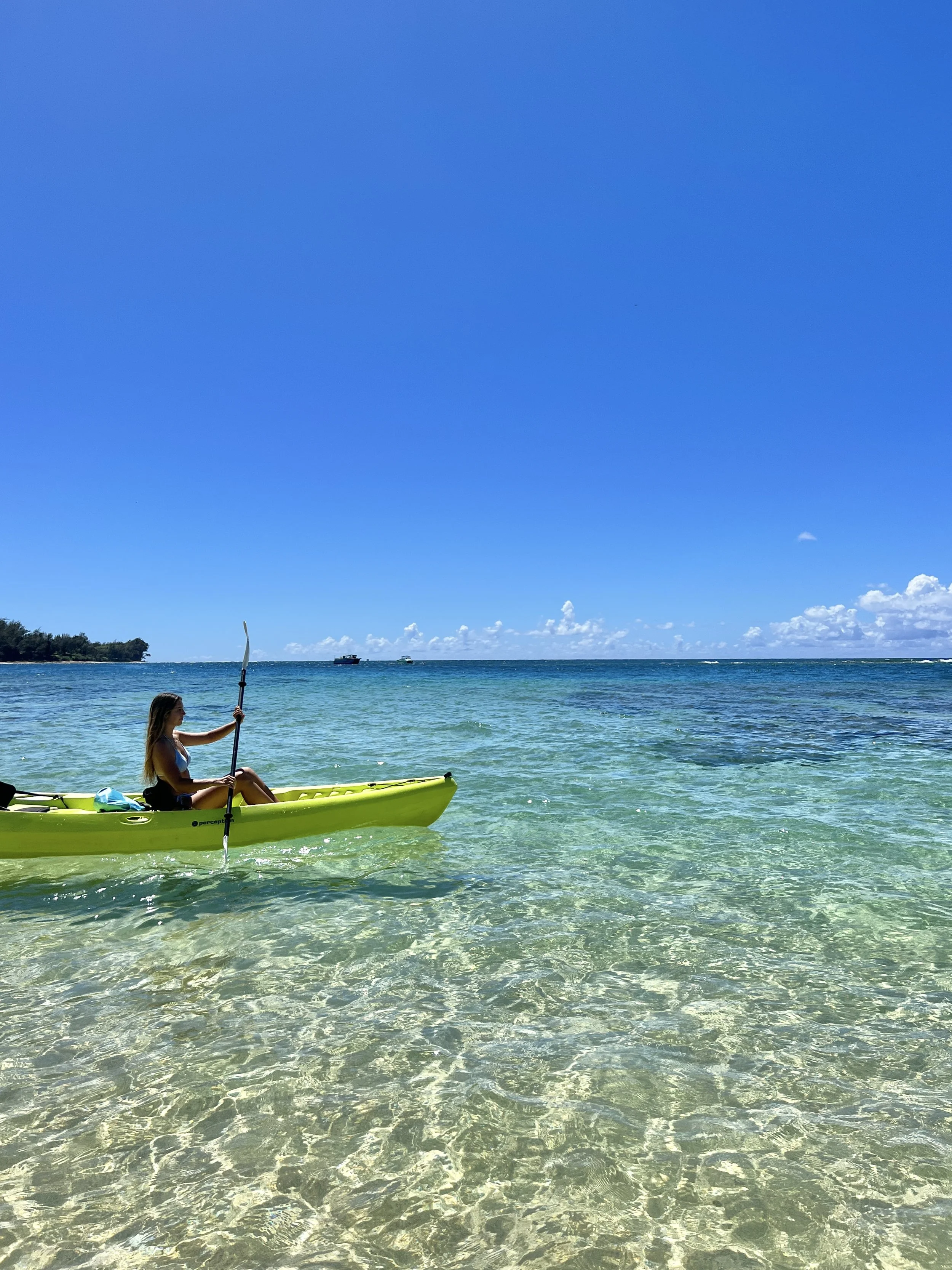 A woman kayaking in clear, shallow ocean water under a bright blue sky.