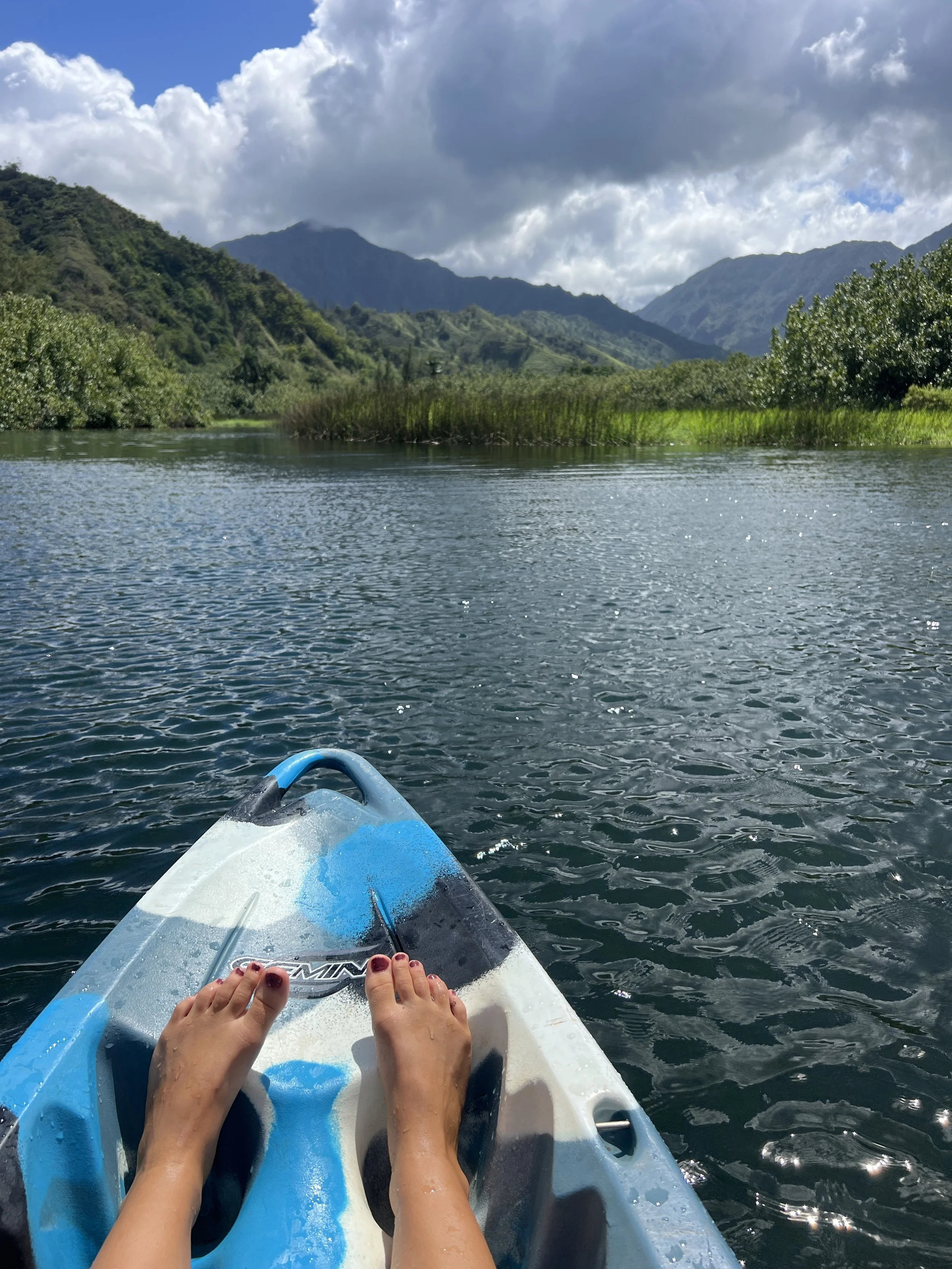 Person's feet on a blue and white kayak paddleboarding on a calm river with green mountains and partly cloudy sky in the background.