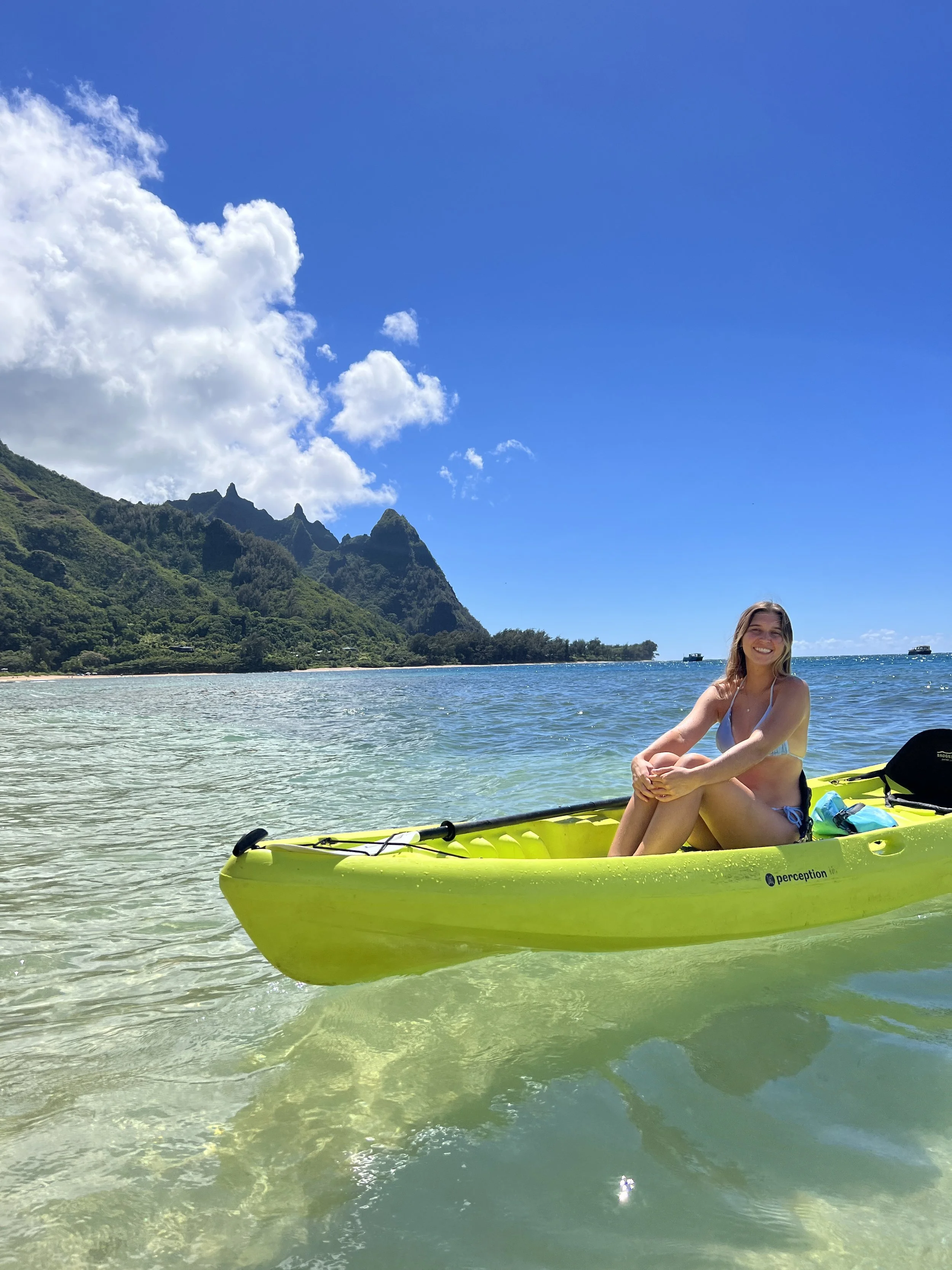 Woman sitting in a yellow kayak on a clear ocean with green mountains and a blue sky in the background.