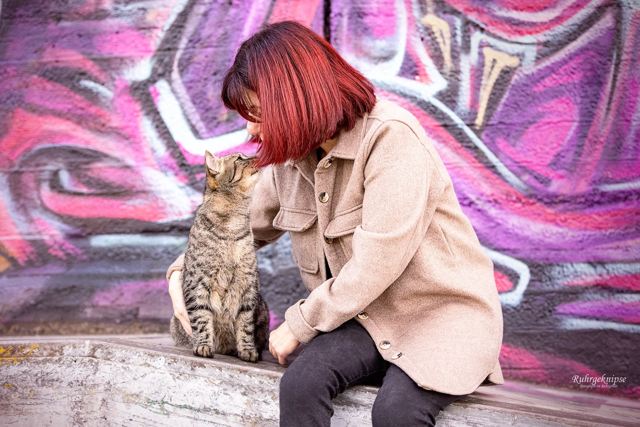 Frau mit roten Haaren knuddelt eine streichelnde Katze vor einer bunten Graffiti-Wand.
