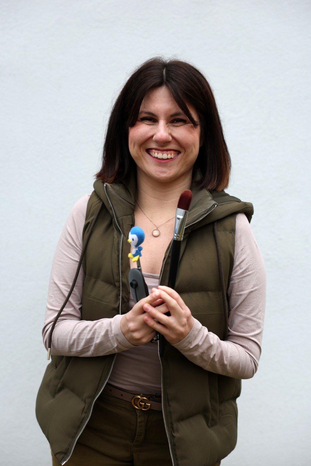Eine lachende Frau hält mehrere Malpinsel in den Händen. Sie trägt eine beige langärmlige Bluse, eine grüne Weste und einen Gürtel mit einem GG-Logo. Im Hintergrund eine weiße Wand.